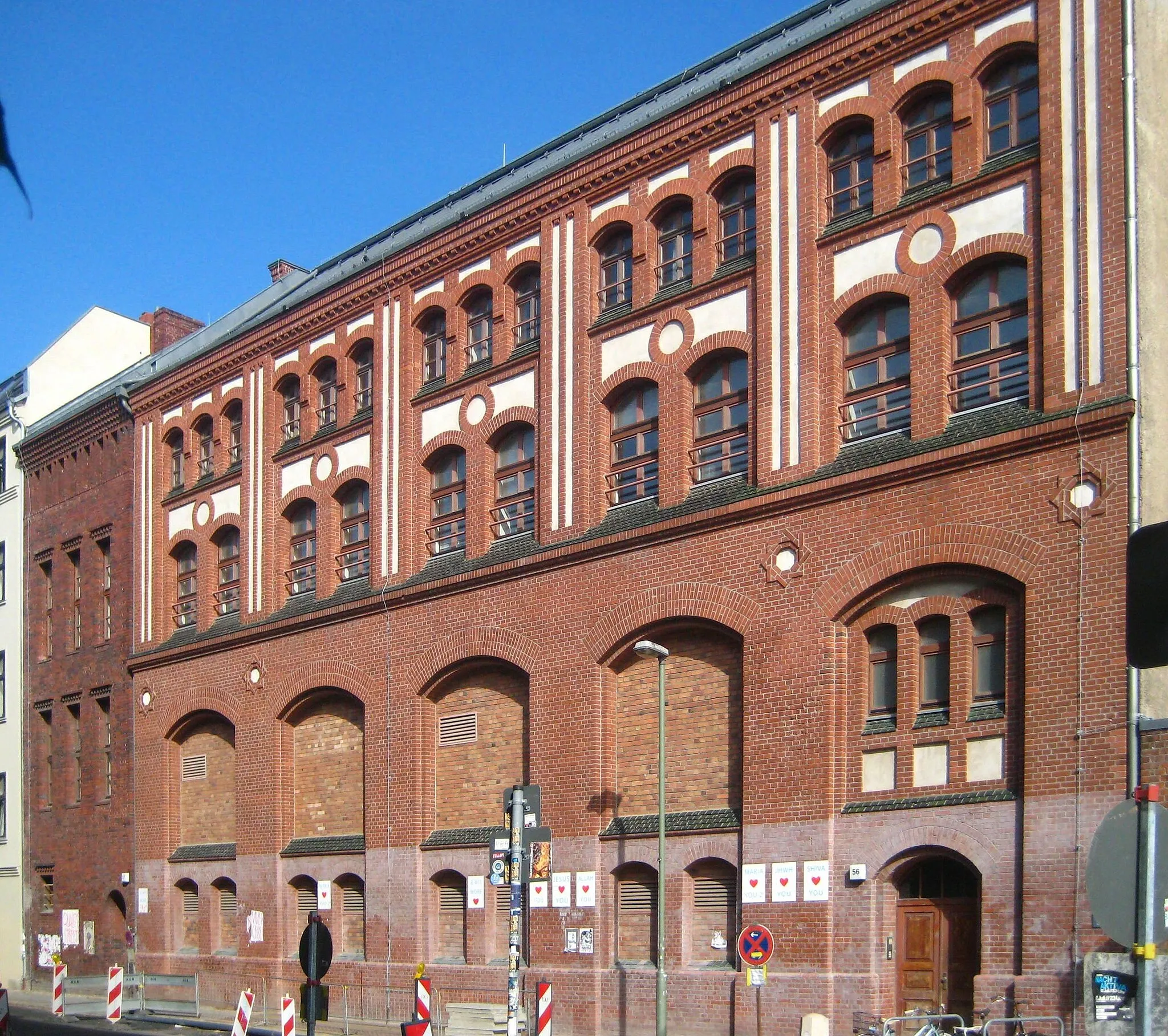 Photo showing: Das ehemalige Abspann-, Umform- und Umspannwerk Koppenplatz in Berlin-Mitte, hier die Fassade an der Auguststraße 56-57. Die 1906 errichtete Anlage diente dazu, den in den Kraftwerken Oberspree und Moabit erzeugten Drehstorm zum Endverbrauch in der inneren und äußeren Spandauer Vorstadt in Gleichstrom umzuformen und die Spannung zu transformieren. Die Gestaltung der repräsentativen Straßenfassaden übernahm der Architekt Franz Schwechten, der dem zwischen Mietshäusern gelegenen Baukörper eine Backsteinfassade gab. Dabei verzichtete Schwechten auf eine einheitliche Form und hob so die technische Funktion des Gebäudes heraus. Die ursprüngliche Raumaufteilung zwischen der hohen Umformerhalle im Erdgeschoss und den niedrigeren Räumen für Transformatoren und Batterien lässt sich daher noch an der Fassade ablesen; die Hochspannungsebene lag im Kellergeschoss. 1926 wurden vom Architekten Hans Müller zwei Kopfbauten für Treppenhäuser und Nebenräume angefügt. Der Bauteil an der Auguststraße ist dabei vierstöckig (hier links), der turmartige Bauteil am Koppenplatz sechstöckig. Dazwischen liegt ein zweistöckiges Verbindungsteil. Auch Müller wählte Backstein für die Fassade, nahm bei deren Gestaltung aber formalen Bezug auf die Stuckfassaden der umgebenden Mietshäuser. Der Gebäudekomplex ist als Baudenkmal gelistet.