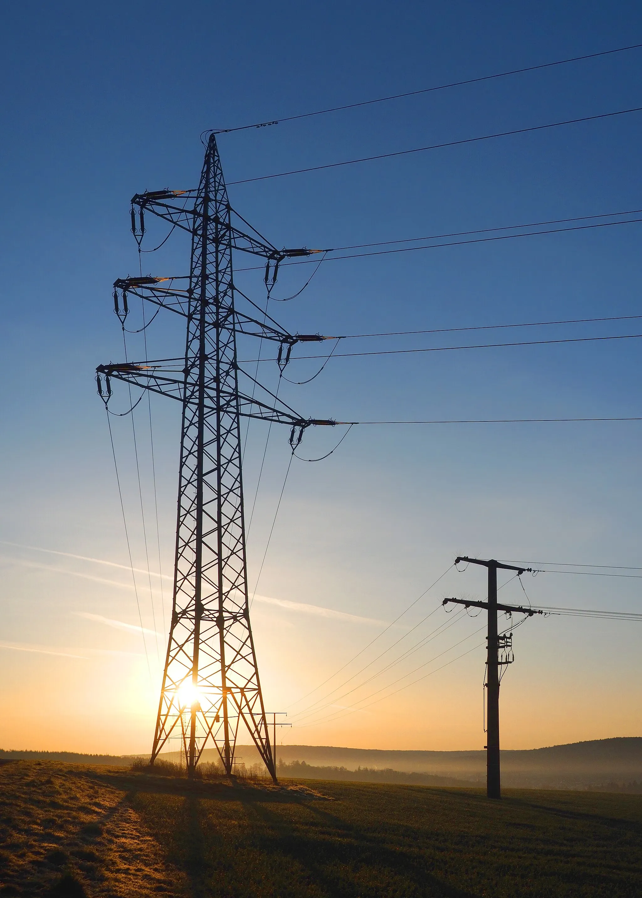 Photo showing: Powerline pylons in Taunusstein, Germany. To the left, 2 circuits of 110 kV 3-phase; to the right, 2 circuits of middle voltage 3-phase (probably 20 kV) with switchable tap. Photo taken 15 minutes after sunrise.