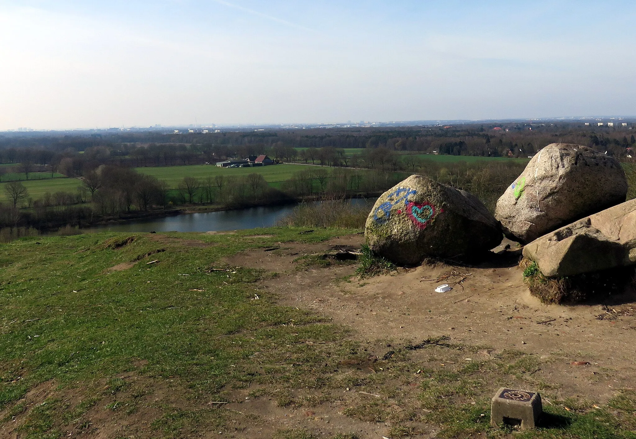 Photo showing: Blick vom Müllberg Hummelsbüttel nach Süden auf den Hummelsee und die Hummelsbütteler Feldmark, vorn Vermessungspunkt, im Hintergrund Wohnwirtschaftsgebäude Glashütter Landstraße 182, bei maximaler Vergrößerung am oberen Waldrand der Flughafen Fuhlsbüttel mit rotem Tower.
This is a photograph of an architectural monument. It is on the list of cultural monuments of Hamburg, no. 26907.