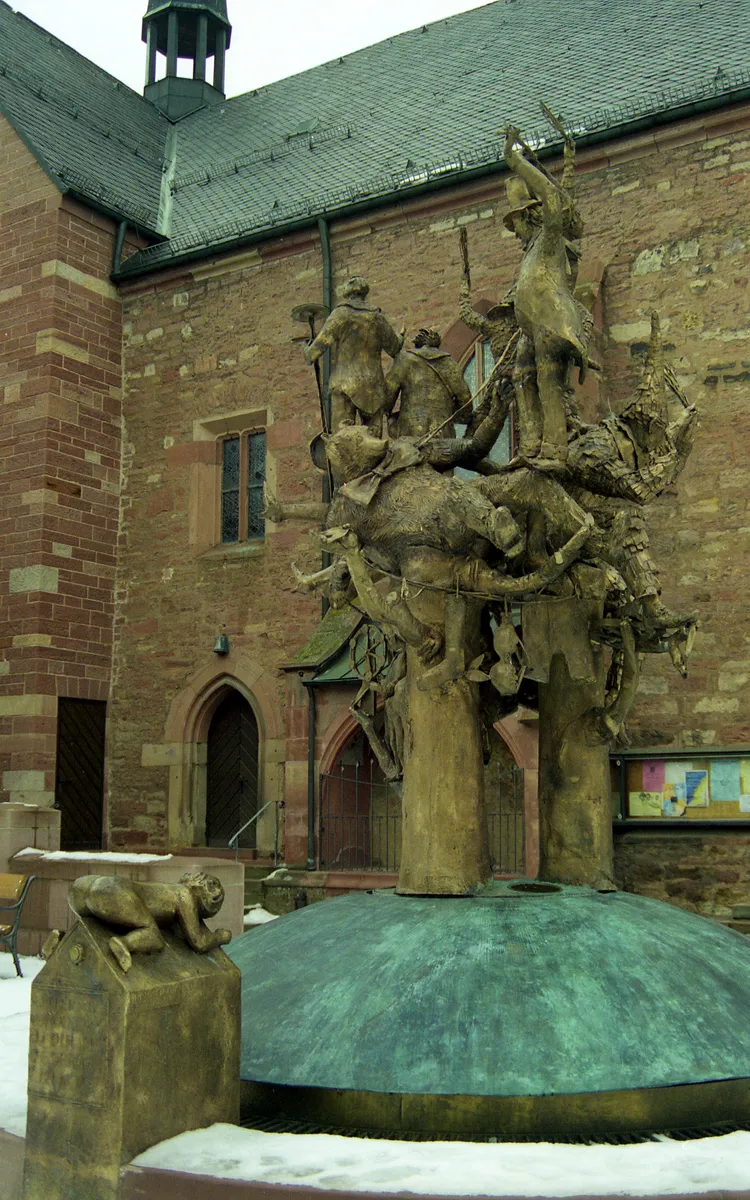 Photo showing: The Narrenbrunnen statue in the Marketplace in Buchen (Neckar-Odenwald-Kreis, Baden-Württemberg), Germany showing various characters of the Buchener Faschenacht (Buchen carnival) above a fountain representing three wells (turned off for winter).
The characters include the Erbsenstrohbär (pea-straw bear) and the Huddelbätz, with pointed hats and costumed in strips of coloured rags or ribbons, who wear bells and make loud noises to drive out evil spirits.
In the foreground is the legendary figure of the "Blecker", who dates from a time when Buchen was beseiged. As their food began to run out, the inhabitants decided to give it all to one man. Once he was fattened up, they displayed him, naked, on top of the walls. The sight of such a fat rump persuaded the attackers that the townspeople obviously still had plenty of food in reserve, so they gave up and retreated.

The sculpture by Joseph Neustifter was unveiled on 11 11 1998, on which occasion the wells were filled with red wine.