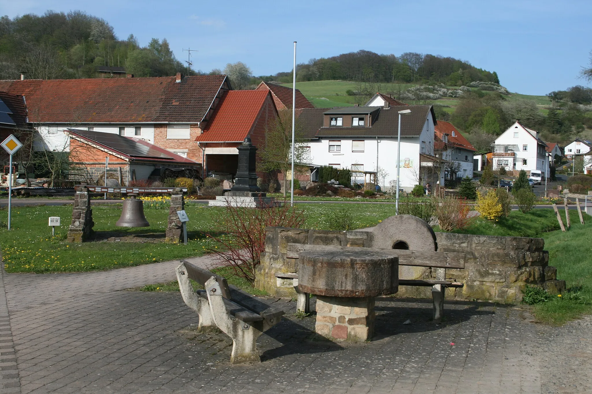 Photo showing: Alter Mühlstein in der Braunhäuser Straße in Gilfershausen. Blick auf Kriegerdenkmal und Alte Glocke.