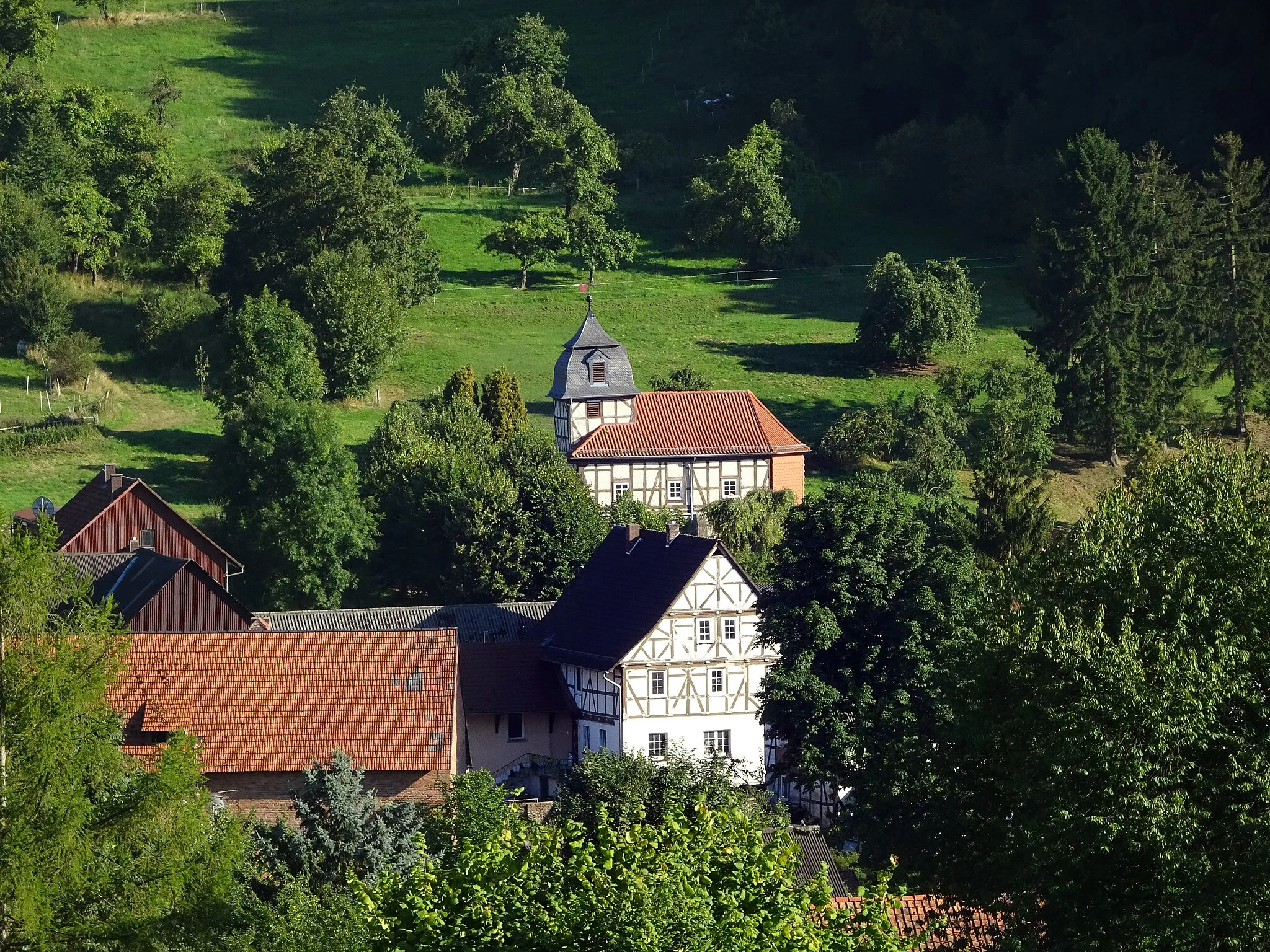 Photo showing: Mönchhosbach, Gemeinde Nentershausen im Landkreis Hersfeld-Rotenburg im Nordosten Hessens.
Die kleine Dorfkirche am Ortsrand von Mönchhosbach stammt in ihrer heutigen Gestalt aus dem Jahr 1781. Eine Inschrift an der Nordseite der Kirche zeugt davon. Vermutlich stand schon vorher eine Kirche an der gleichen Stelle, von der Mauerwerk und Fundamente beim Neubau wieder verwendet wurden. Die heutige Kirche besteht aus zwei Geschossen. Das Erdgeschoss ist aus Bruchstein gemauert, das obere in Fachwerk gezimmert. Dieser Fachwerkausführung verdankt die Mönchhosbacher Kirche ihre Aufnahme in die Reihe der förderungswürdigen Fachwerkkirchen in Hessen und damit einer Grundrenovierung und Restaurierung in den Jahren 1980 bis 1983.