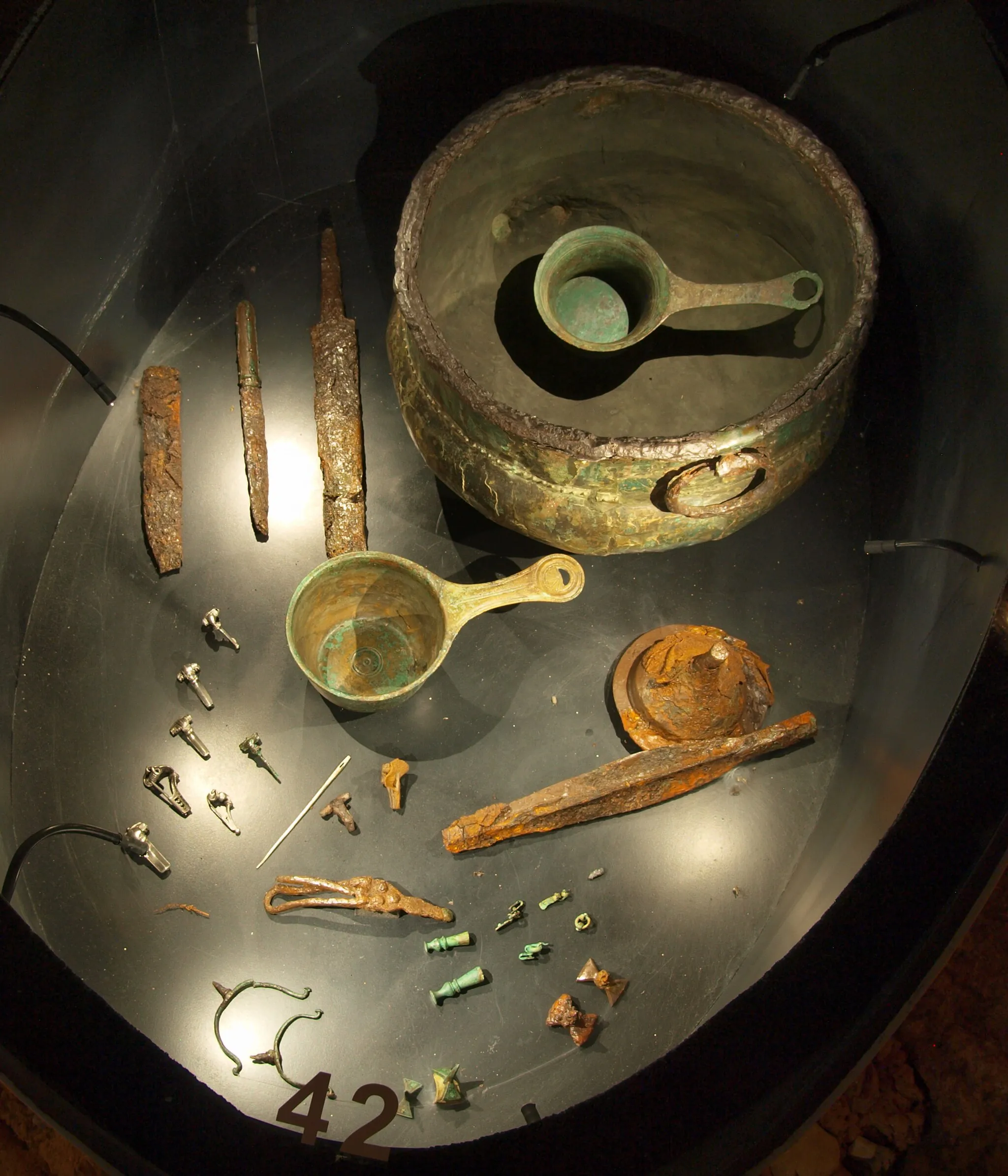 Photo showing: Items from the Lombard warrior's grave Putensen 150, dating to circa 50 A.D., in the community Salzhausen, District of Harburg, Germany. It consists of a Roman bronze cauldron, 2 Roman casseroles (patherae), sword, dagger, knife, 8 fibulae, silver pin, shield boss, lance point, belt buckle, 2 drinking horn fittings, 3 pairs of spurs. Photographed at Archaeological Museum Hamburg, Germany.