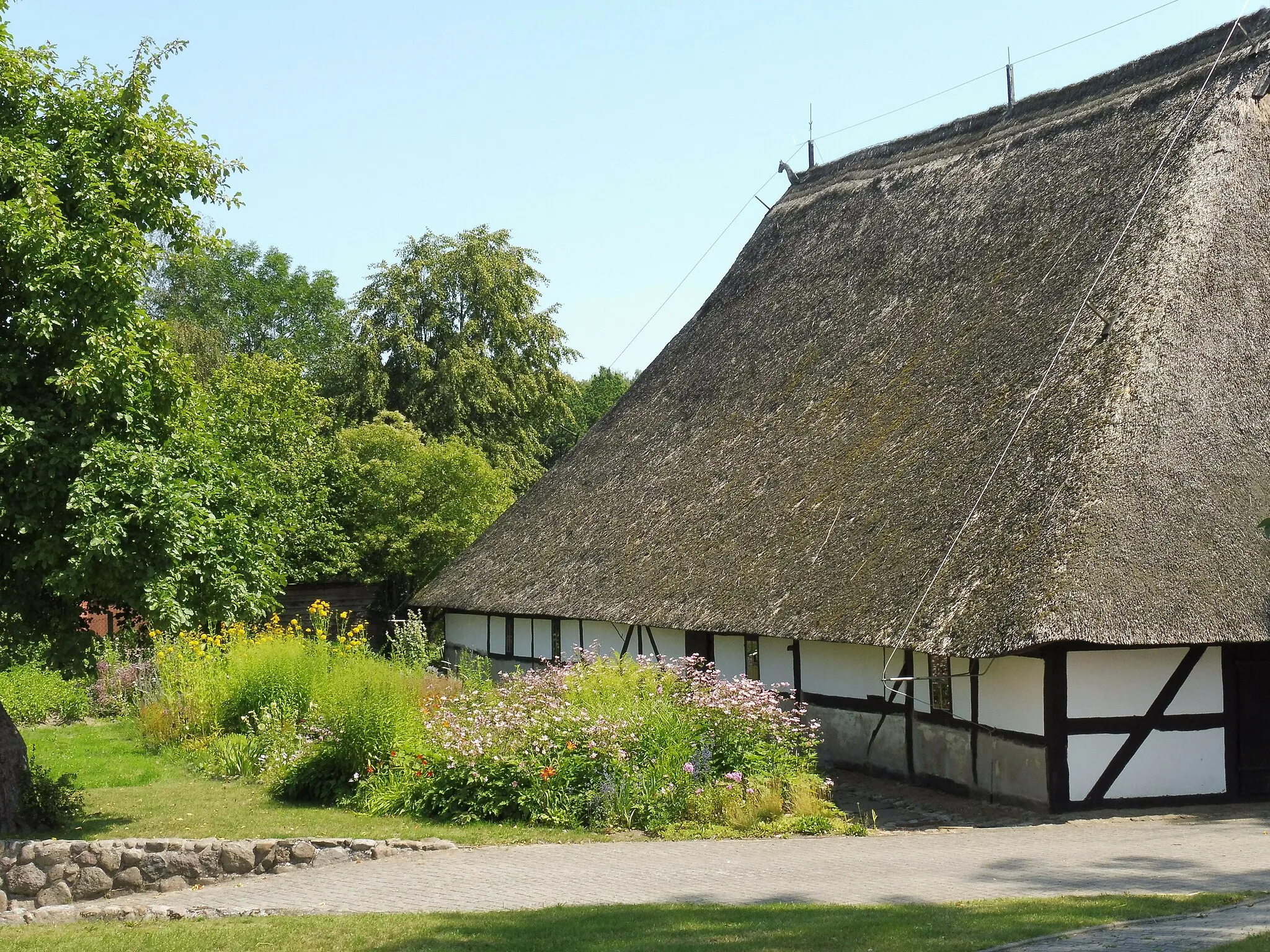 Photo showing: This Low German house is listed. It is located Dorfstrasse 8 in Schwerin in Mecklenburg-Vorpommern and home to family-owned village museum Dat oll' Hus (Low German for The old house). Side view shows the vast roof that sweeps down to just above head height. Next to the side is a typical bed of a cottage garden.