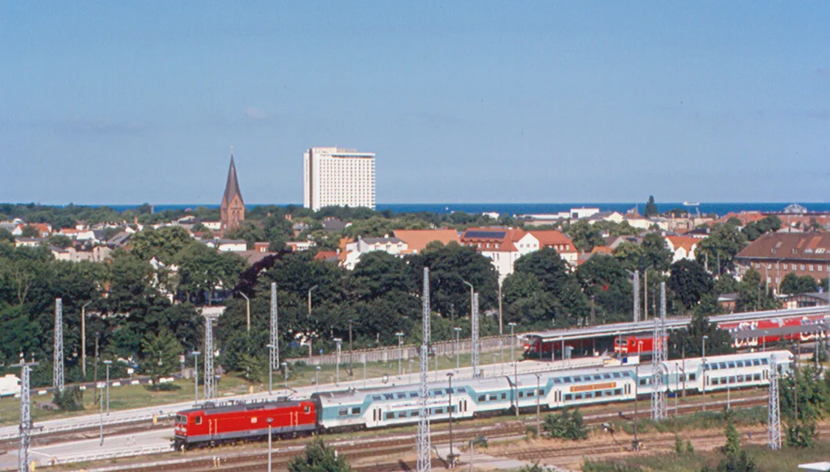 Photo showing: Warnemünde and the station area from the ship.  This was the port for Rostock, and perhaps one of its more elegant suburbs.  Rostock is about 10 km (6 miles) away.  This was the first I had seen double-decker cars in the Deutsche Bahn (Deutsche Bundesbahn).  Hotel Neptun, which looks like a soviet-style hotel, is in the background.  About half of the ship's passengers had gone to Berlin by special train from the station.  This photo is geotagged.