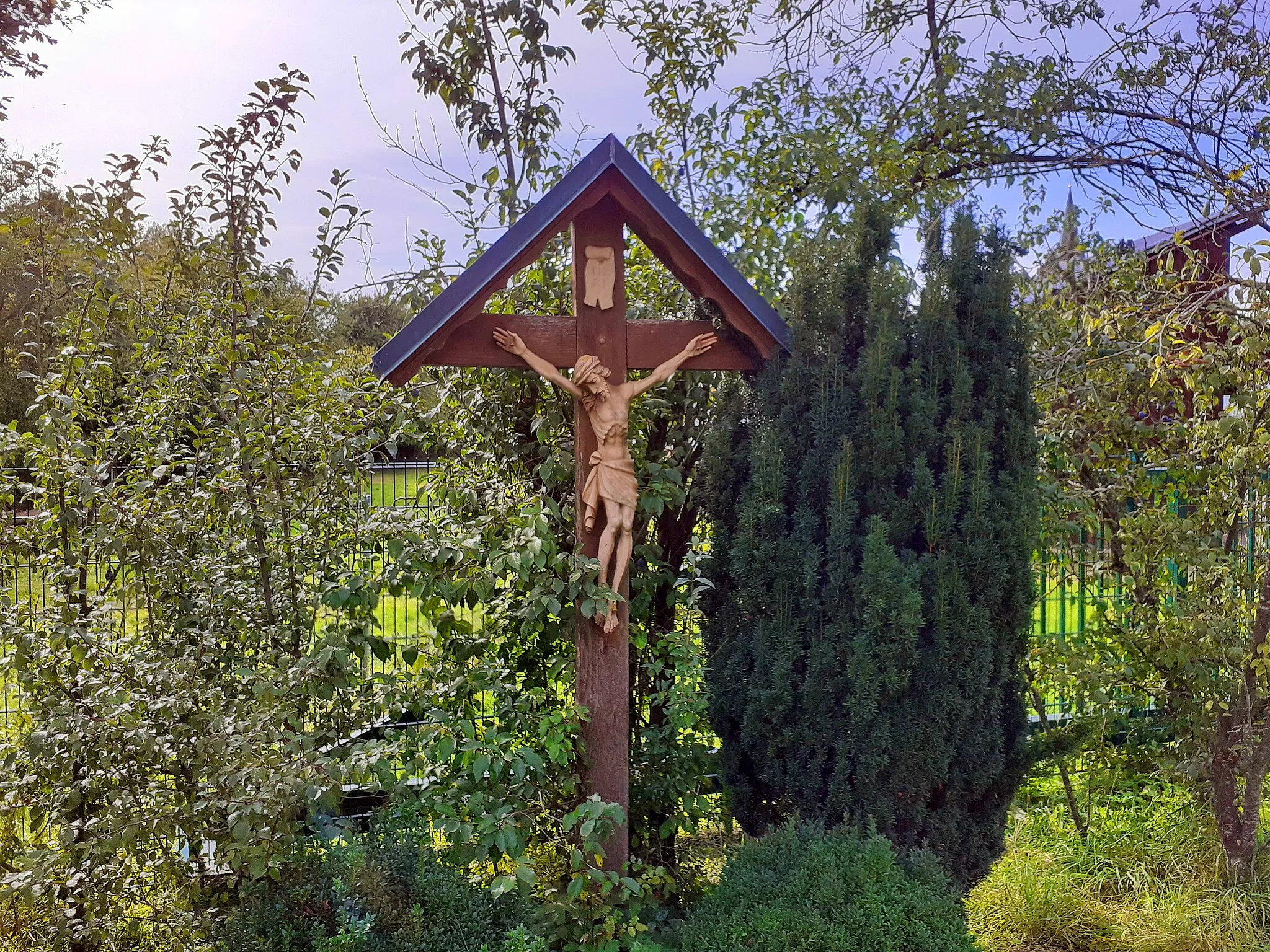 Photo showing: Bei dem Holzkreuz in Moosthann in der Gemeinde Postau handelt es sich um die in der Gegend weit verbreitete Version, die aus Laden gezimmert und mit einem rechteckigen Satteldach gedeckt ist. Das Dach, mit geschweiften Giebelleisten ist zum Schutz vor der Witterung mit Blech verkleidet. Die Rückseite ist nicht verkleidet. Das Kreuz ist nach Osten hin zur Straße ausgerichtet.
Die Figur des Gekreuzigten ist sehr schlank, um nicht zu sagen ausgehungert, dargestellt und mit vier Nägeln an die Kreuzbalken geheftet. Die Nägel der Hände befinden sich in den Handflächen. Das Haupt ist geneigt. Wie üblich befindet sich die oben eingerollte Titulusbanderole INRI am oberen Ende des Kreuzbalkens.

Das Kreuz steht an der Ostseite des Anwesens in der Paindlkofenerstraße 1 vor dem Zaun in einer Sträuchergruppe. Im Hintergrund ist über den Dächern der Kirchturm der Filialkirche St. Jakob von Moosthann zu sehen.