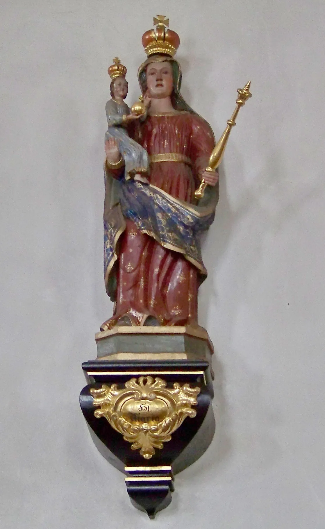 Photo showing: Kirchroth, Dekan-Seitz-Straße 5. Katholische Pfarrkirche St. Vitus, errichtet in der Mitte des 17. Jahrhunderts. An der Nordwand des Langhauses steht eine Madonnenfigur mit Kind auf einer Wandkonsole. Maria trägt das bekleidete Jesuskind im rechten Arm. In der linken Hand hält Maria ein goldenes Zepter. Beide sind mit Kronen in den Farben Rot und Gold bekrönt. Interessant sind die Farbgebung und das Muster von Marias rotem Kleid und dem blauen Schleier.