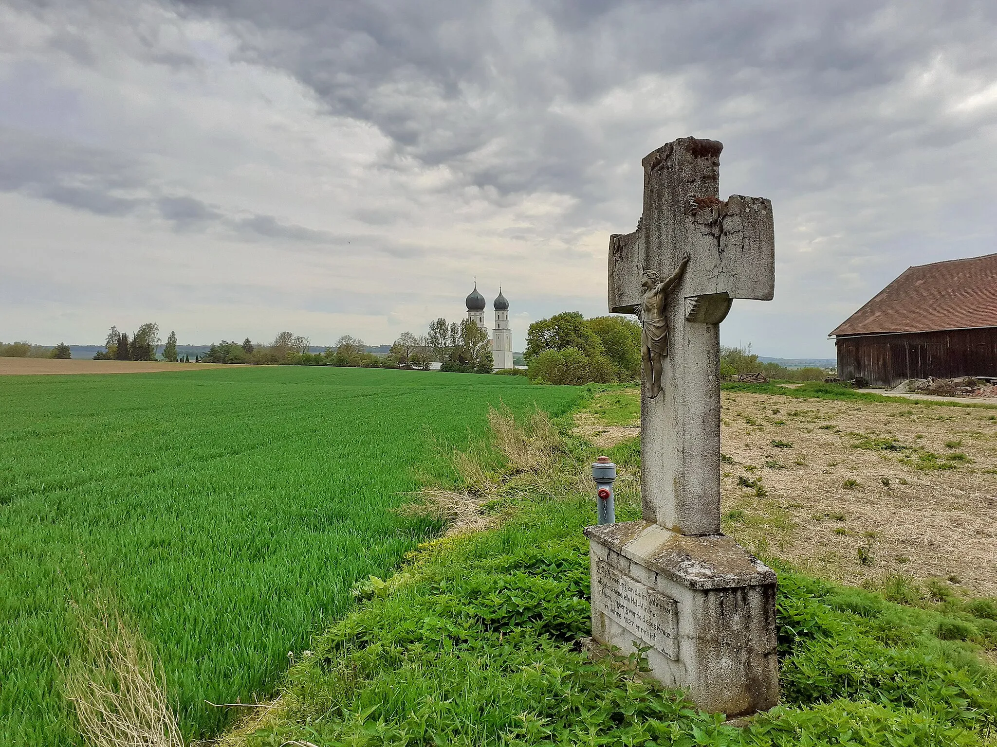 Photo showing: Haindlingberg gehört zur Stadt Geiselhöring und liegt etwa 350 Meter nördlich des Wallfahrtsorts Haindling. Das massige Steinkreuz steht südöstlich des Einödhofes. Auf einem Sockel, dem ein breiter Quaderstein folgt, auf den eine flache pyramidenförmige Platte aufgelegt ist, erhebt sich das Kreuz mit verbreiteten Enden. Der metallene Korpus entspricht dem Dreinageltypus.
Bis 1924 stand am Hof eine stattliche Barockkirche aus dem Jahre 1730 deren Turm 1790 erhöht wurde. 1924 wurde die Kirche baufällig und abgebrochen. Zur Erinnerung wurde das Kreuz aufgestellt. Nach einem Beschluss des Konzils von Trient (1545 - 1563) war auf dem Platz einer abgebrochenen Kirche ein Steinkreuz zu errichten.
Die Kirche mit dem Patrozinium Stefan und Michael hatte einen eingezogenen, halbrunden Chor mit einer Fensterachse und Kreuzgratgewölbe. Das Schiff mit Flachdecke erstreckte sich über drei Fensterachsen. Der Turm mit Spitzhelm über vier Giebeln befand sich nördlich auf Höhe des Chorbogens. Die durchaus kostbare figurliche Ausstattung befindet sich heute in der nahen Wallfahrtskirche in Haindling.
Eine Schrifttafel am Sockel berichtet über den Grund der Errichtung des Kreuzes:
"Zur Erinnerung an die im Jahre 1924 abgebrochene alte Hof-Kirche in Haindlingberg wurde dieses Kreuz im Jahre 1927 errichtet."

Vom Kreuz aus sieht man die südlich stehenden Türme der Wallfahrts- und Kreuzkirche von Haindling.