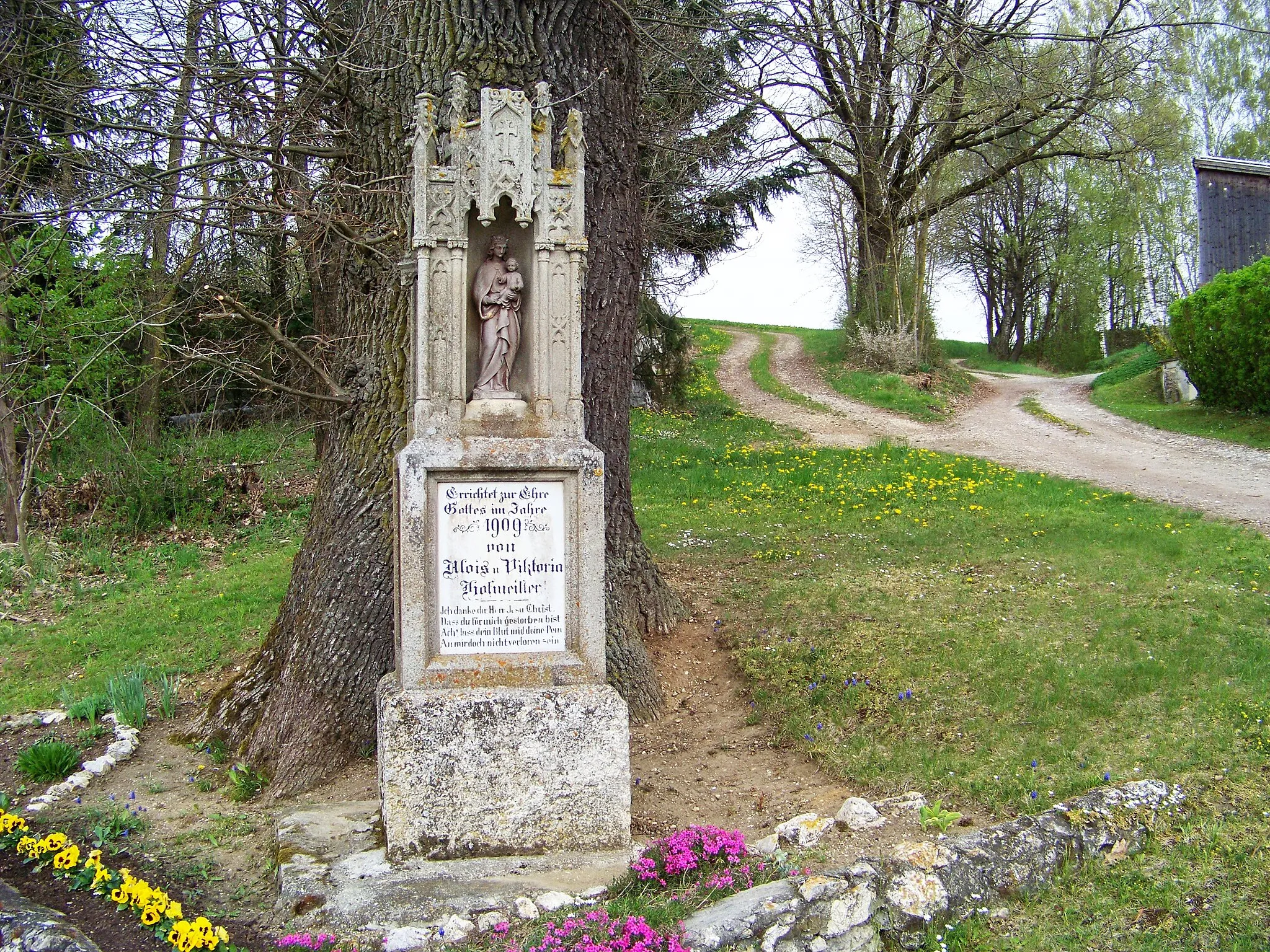 Photo showing: In Hadersbach steht die Kreuzsäule östlich der SR53 zwischen den Hausnummern 6 und 10 vor einer mächtigen Eiche. Der Vorplatz ist mit einer Mauer aus Kalk-Bruchsteinen eingefasst um mit einem Blumenbeet geschmückt. Die Kreuzsäule enthält auf drei Seiten Nischen mit Figuren und drei Schrifttafeln. Früher war die Säule mit einem Kreuz bekrönt, das nicht mehr vorhanden ist.
Einem nach oben abgefasten Quadersockel aus Granit folgt ein Steinblock, der auf drei Seiten Schrifttafeln enthält. Darauf steht der Aufsatz aus Sandstein mit drei Figurennischen, die mit Ornamenten und Fialen im Stil der Neugotik verziert sind. An der Vorderseite befindet sich eine Statuette, die Maria mit dem Jesuskind darstellt. Marias Haupt ist bekrönt. Sie trägt das Jesuskind auf dem Arm. Jesus hält in der linken Hand eine Kugel und hat die rechte Hand zum Segen erhoben.
Die Tafel an der Vorderseite trägt folgende Aufschrift:
"Errichtet zur Ehre Gottes im Jahre 1909 von Alois u. Viktoria Hofmeister."
Es folgt das Gebet:
"Ich danke dir Herr Jesu Christ,
Dass du für mich gestorben bist.
Ach lass dein Blut und deine Pein
An mir doch nicht verloren sein."
In der östlichen Nische steht eine Statuette mit einem Buch in der Hand. Möglicherweise handelt es sich um eine Darstellung der heiligen Anna, der Mutter Marias.
Darunter steht am Sockel:
"Gott segne unsere Fluren"
In der westlichen Nische steht eine weitere Figur, die ein kleines Kreuz an ihre Brust drückt. Es könnte sich um die Darstellung der heiligen Theresa vom Kinde Jesu handeln.
Darunter steht auf der Schrifttafel:

"Zur Erinnerung an 1. Mai 1909"