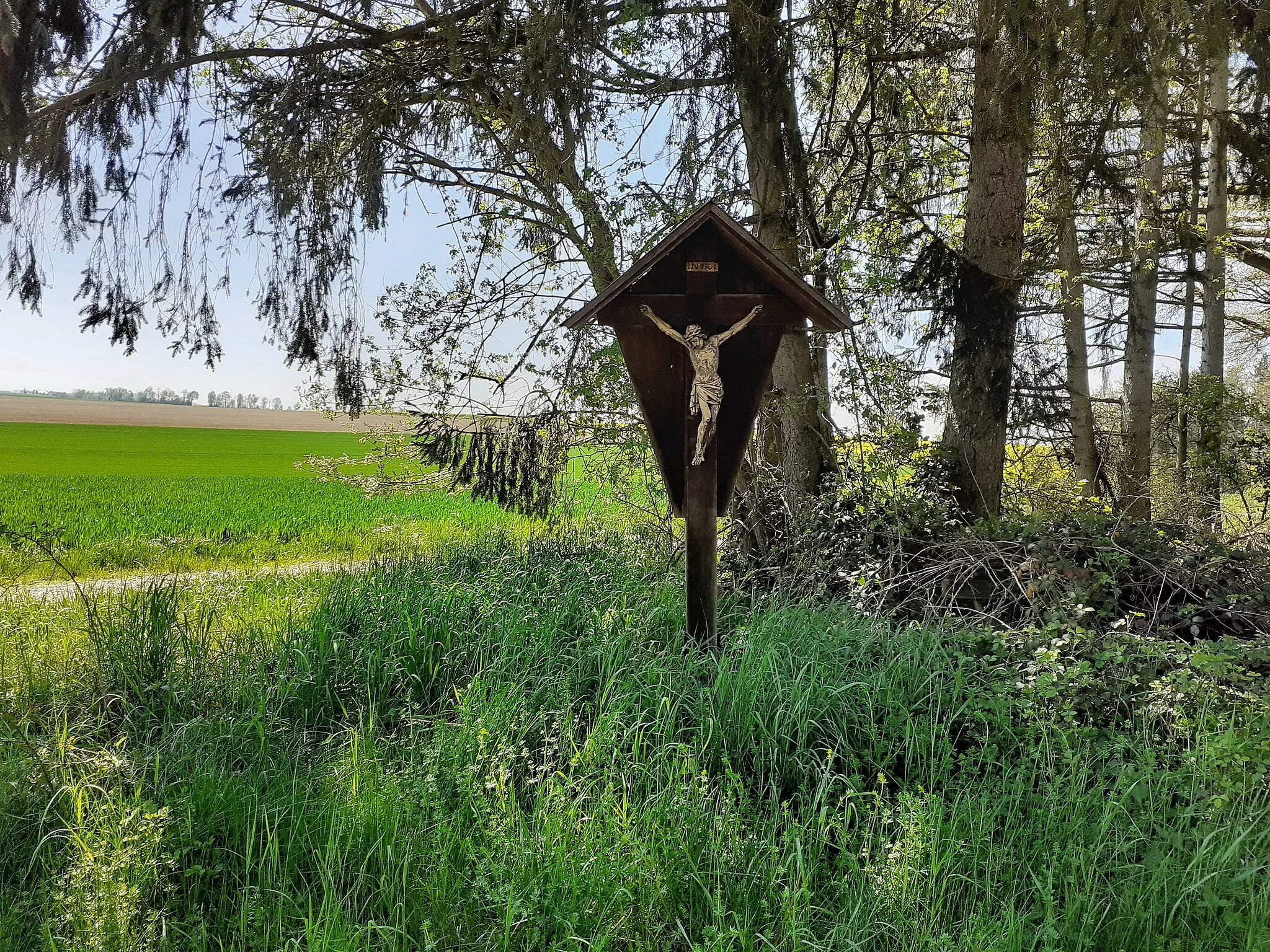 Photo showing: An der SR18 von Metting nach Eschlbach in der Gemeinde Leiblfing steht vor Eschlbach dieses Kreuz an der Westseite der Straße am Waldrand. Der Wald dahinter trägt den Namen "Auf der Heide". Die südlich davor liegenden Flurnamen lauten "Breite" und "Fuchsberg".
Das Kreuz ist aus massiven Kanthölzern gezimmert, rückseitig verbrettert und mit einem breiten, rechtwinkeligen Satteldach, das mit Blech eingeschlagen ist, vor der Witterung geschützt. Die Christusfigur ist mit drei Nägeln befestigt. Der Titulus INRI ist als schmales, rechteckiges Schild gefertigt.