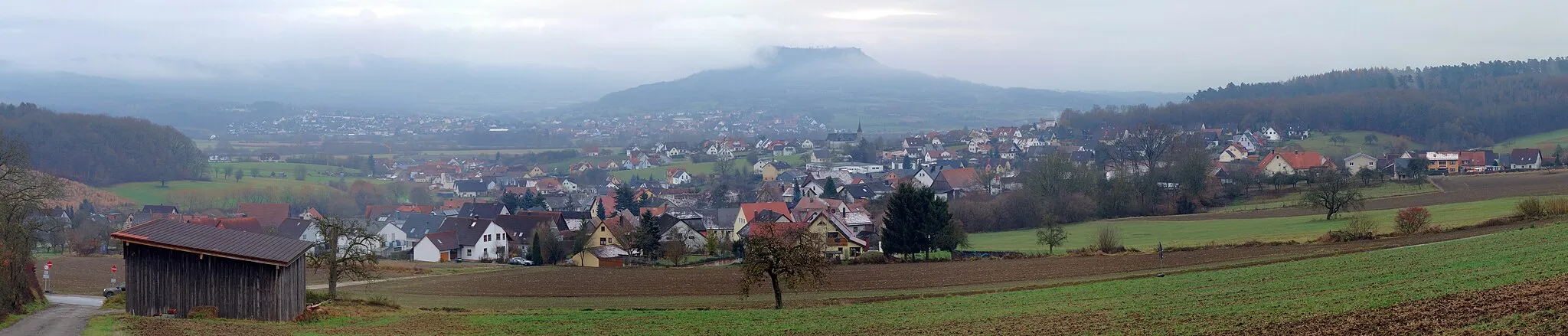 Photo showing: A panorama of Weilersbach, a town in northern Bavaria. In the foreground, its village of Mittlerweilersbach can be seen. Beyond it is its village of Unterweilersbach with its church St. Anna. In the background, partially obscured by clouds the hill Ehrenbürg is visible, and along its slopes the town of Kirchehrenbach can be seen.