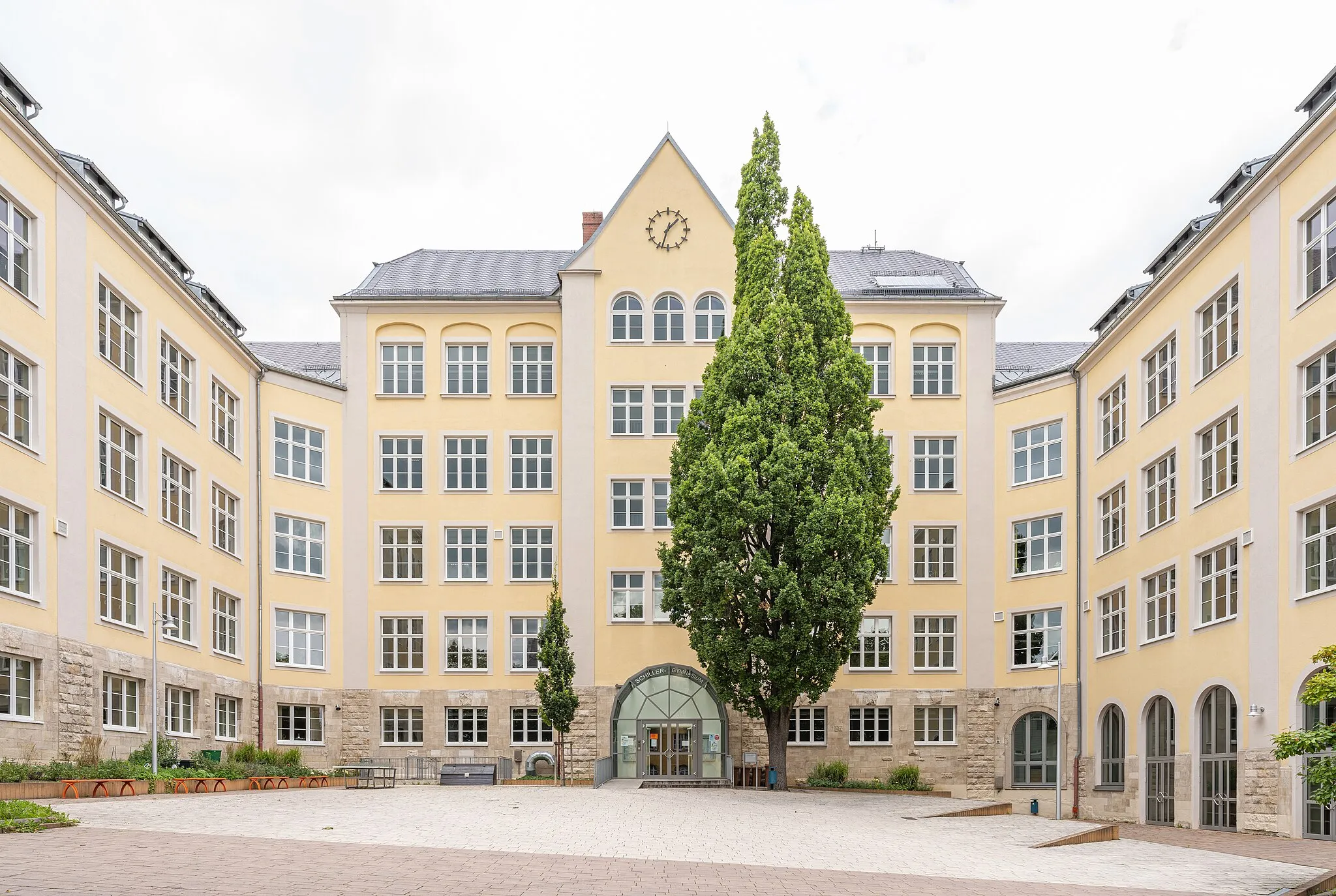 Photo showing: Facade of the main entrance of the Schiller Gymnasium Hof. On the left and right you can see a part of the west and east wing of the school building. In the center of the picture you can see the playground. The glass facade represents the main entrance and bears the lettering of the school name. The building is U-shaped and has an east wing (on the right of the picture) and a west wing (on the left of the picture).