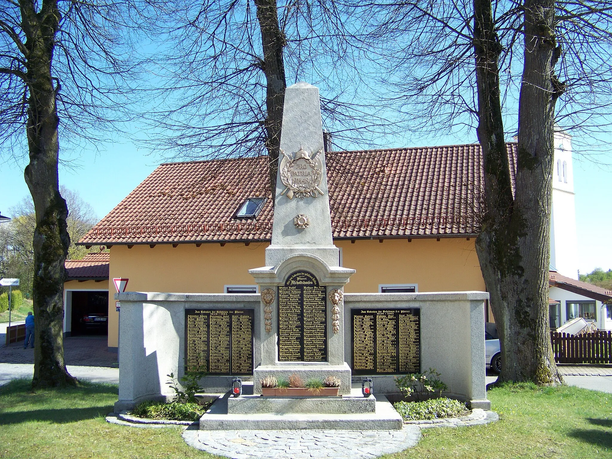 Photo showing: In der dreieckten Verkehrsinsel zwischen der B15 und der Dorfstraße steht das imposante Kriegerdenkmal in einer Baumgruppe. Drei Stufen führen zum Mittelteil. Seitlich sind Blumenbeete angepflanzt. Auf drei großen Tafeln sind die Namen der Gefallenen der beiden Weltkriege in goldenen Buchstaben eingemeißelt. Im Mittelteil erhebt sich ein hoher Obelisk, den ein Relief mit Eichenlaub, Fahnen und Helm ziert. Darauf ist der heroische Spruch zu lesen: PRO PATRIA 1914-18. (für das Vaterland) Unterhalb ist ein Malteserkreuz angebracht. Zu beiden Seiten der mittleren Tafel, auf der die Gefallenen des ersten Weltkriegs aufgeführt sind, befinden sich  Reliefs mit Löwenköpfen und Fruchtgehängen. Erschütternd ist zu lesen welch große Zahl an jungen Männern ihr Leben in den europäischen Nachbarländern verloren haben.