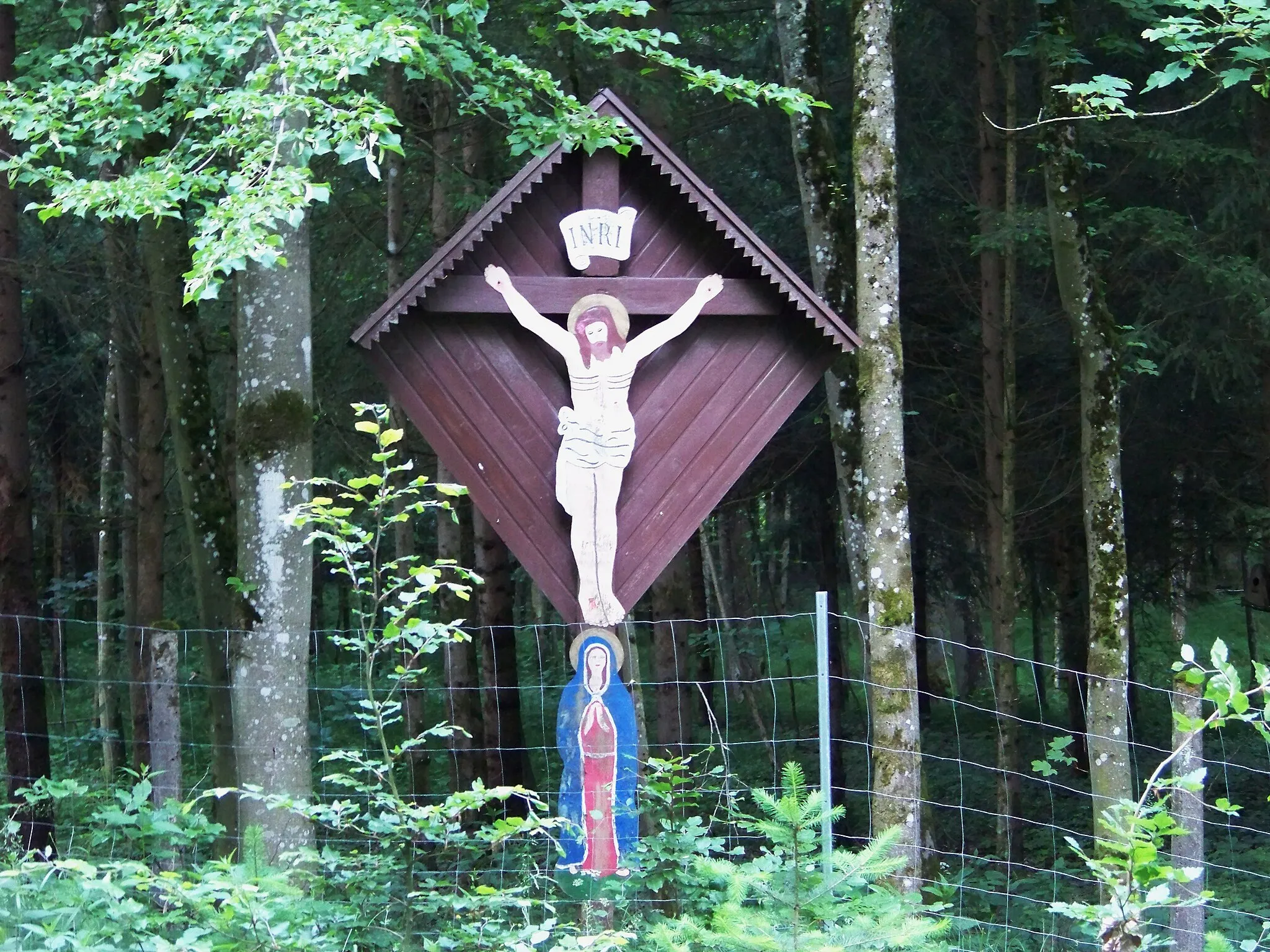 Photo showing: In der Gemeinde Laberweinting, östlich von Asbach, am Wirtschaftsweg zwischen Buchetholz und Brunnberg, der zur Kiesgrube Steimer führt, steht das Kreuz rechts neben der Straße in einem umfriedeten Waldstück in der Nähe eines Quellbaches des Flüsschens Asbach.
Das große Kastenkreuz im Brunnberg ist auf der Rückseite rautenförmig verbrettert. Die Bedachung ist mit Blech verkleidet und an der Vorderseite gezahnt. Am Holzkreuz ist ein polychromer Blechschnitt-Corpus-Christi mit Gloriole, Dornenkrone und Lendenschurz befestigt. Über dem Gekreuzigten eine Blechfahne mit den Initialien INRI. Unterhalb ist ein polychromes Blechbild der schmerzhaften Mutter Gottes angebracht.