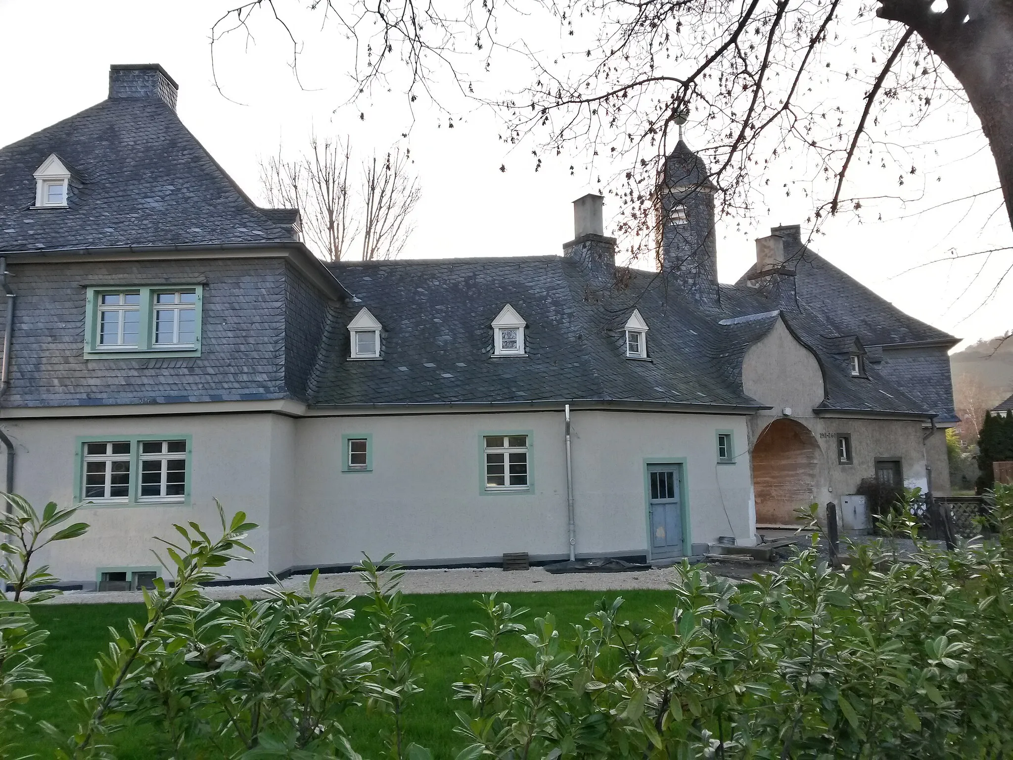 Photo showing: Himmelsbachsiedlung in Kempten am Rhein (Bingen am Rhein),  Mainzer Straße 242–260: kleine Arbeitersiedlung der Himmelsbachwerke, geschlossenes Polygon mit fünf Doppelhäusern in Heimatstilformen, Architekt Prof. Cäsar, Karlsruhe. Straßenseitiger Torbogen durch geschweiften Giebel und glockenturmartigen Aufsatz akzentuiert. Die Haupthäuser
sind durch ihre Nebengebäude untereinander verbunden,
durch weitere kleine Torbögen gelangt man in die umgebenden

Kleingärten.