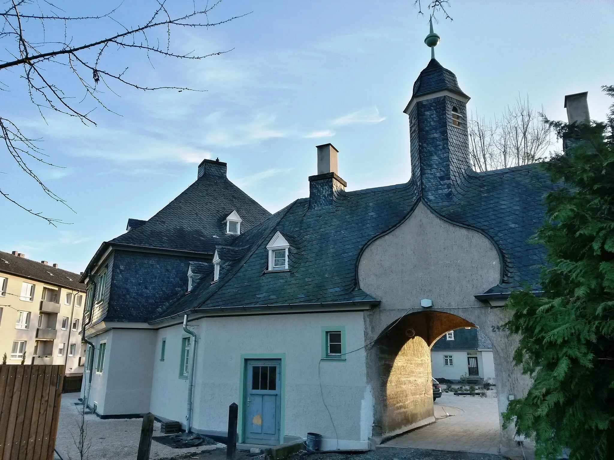 Photo showing: Himmelsbachsiedlung in Kempten am Rhein (Bingen am Rhein),  Mainzer Straße 242–260: kleine Arbeitersiedlung der Himmelsbachwerke, geschlossenes Polygon mit fünf Doppelhäusern in Heimatstilformen, Architekt Prof. Cäsar, Karlsruhe. Straßenseitiger Torbogen durch geschweiften Giebel und glockenturmartigen Aufsatz akzentuiert. Die Haupthäuser
sind durch ihre Nebengebäude untereinander verbunden,
durch weitere kleine Torbögen gelangt man in die umgebenden

Kleingärten.