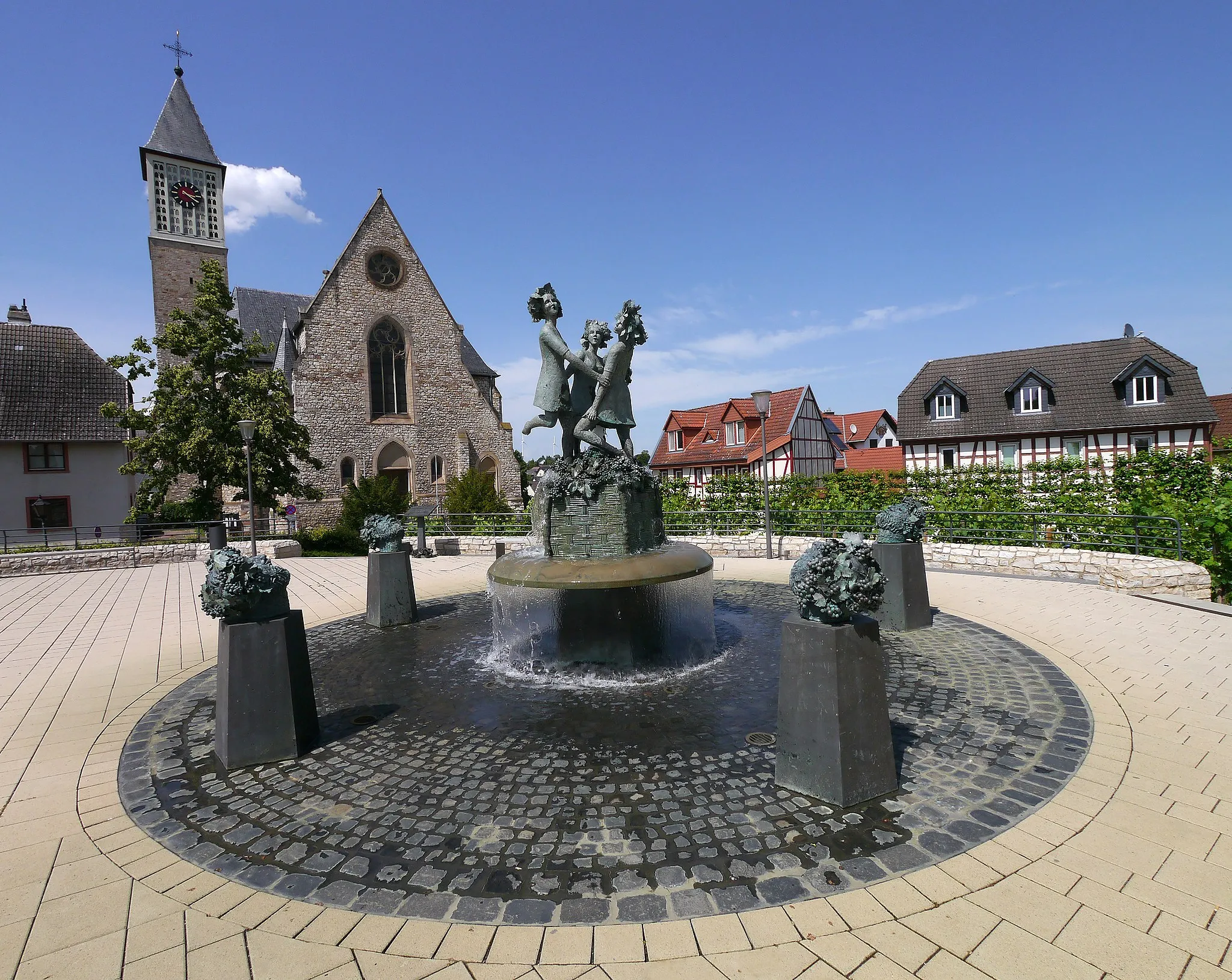 Photo showing: Drei Grazien Brunnen auf dem Linden-Platz in Zornheim vor der Kirche St. Bartholomäus (2021). Zum Brunnen: "Der Brunnen wurde von der Familie Peter Eckes gestiftet und von dem Bildhauer Franz Müller-Steinfurth gestaltet und realisiert. Drei Grazien treten Trauben in einer fünfeckigen Kelter zu Most. Sie werden dabei von fünf Bacchanten beäugt. Diese symbolisieren die fünf Weinlagen Zornheims: Dachgewann, Guldenmorgen, Mönchbäumchen, Pilgerweg und Vogelsang. Tag der Einweihung 26. April 2015. Text der Infotafel entnommen."