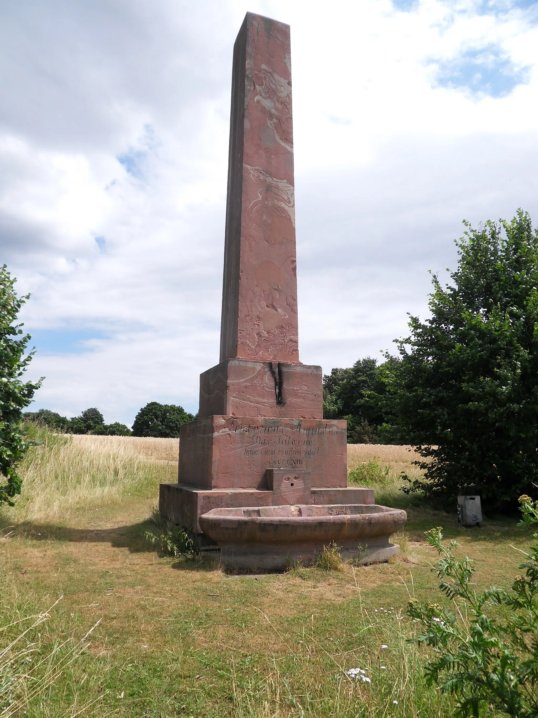 Photo showing: Das Wandersmann-Denkmal von 1813 am Wiesbadener Kreuz über der Bundesautobahn 66 bei Hofheim-Wallau, Ansicht von Westen. Es handelt sich um einen Sandstein-Obelisk mit dreistufigem Sockel Die Inschrift in lateinischer Sprache auf dem Sockel erinnert an die Fertigstellung der Chaussee, deren Verlauf heute die Autobahn folgt, und an den Bauherrn, Herzog Friedrich August von Nassau. Ursprünglich handelte es sich um ein Brunnendenkmal mit einem bronzenen Löwenkopf als Wasserspeier und einem Brunnentrog. Der Löwenkopf ist in den letzten Jahren durch Vandalismus abhanden gekommen. Er wurde aus seiner Verankerung heraus gebrochen. Seine Umrisse sind an der helleren Farbe des Sandsteins erkennbar.