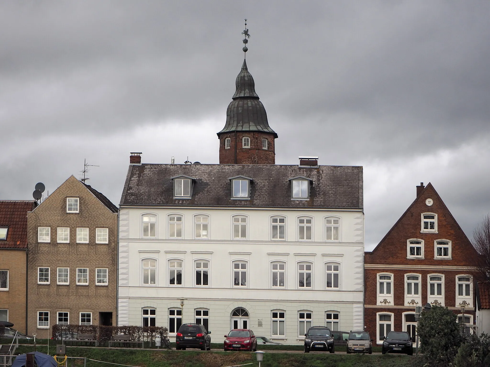 Photo showing: Glückstadt (Schleswig-Holstein) – Am Hafen 40 – Königshof mit Wiebke-Kruse-Turm – Baujahre: 1873/74 1630/31 – Der Königshof“ war das  erste Wohnhaus König Christians IV. aus der Gründungszeit der Stadt. Er nutzte es bis zur Fertigstellung seines Glückstädter Schlosses, das bis 1631 in unmittelbarer Nähe errichtet wurde. 1638 ging das Palais als Geschenk an seine Geliebte Wiebke Kruse. Das Gebäude wurde nach einem Brand im 19. Jahrhundert abgebrochen. Erhalten blieben der polygonale Treppenturm mit geschweifter Doppelhaube, Reichsapfel und Reiter an der Wetterfahnenstange sowie Wendeltreppe und der gewölbte Keller.

– Auf den Fundamenten des abgebrochenen Gebäudes wurde bis 1873/74 ein Wohngebäude in den Formen des Historismus errichtet (weißes Haus im Bild). Es ist dreigeschossiger Traufenbau mit dezenter spätklassizistischer Putzgliederung und dekorativ ausgemaltem Eingangsflur.