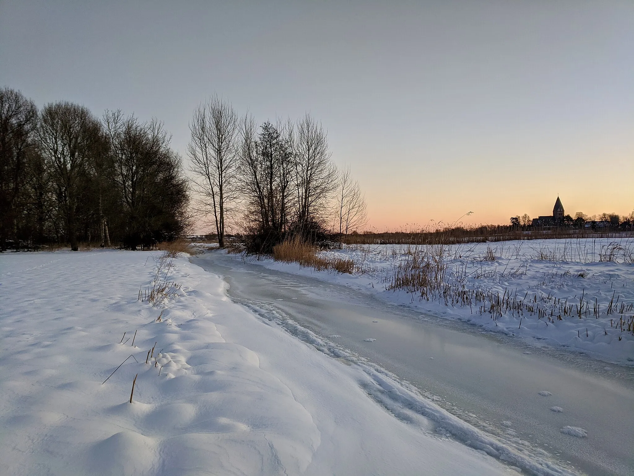 Photo showing: View over the meadows of the Kremper Au stream between Logeberg and Altenkrempe. Snow covers the meadows, the side arm of the Kremper Au is frozen to ice. The chuch in the background is the Basilika Altenkrempe