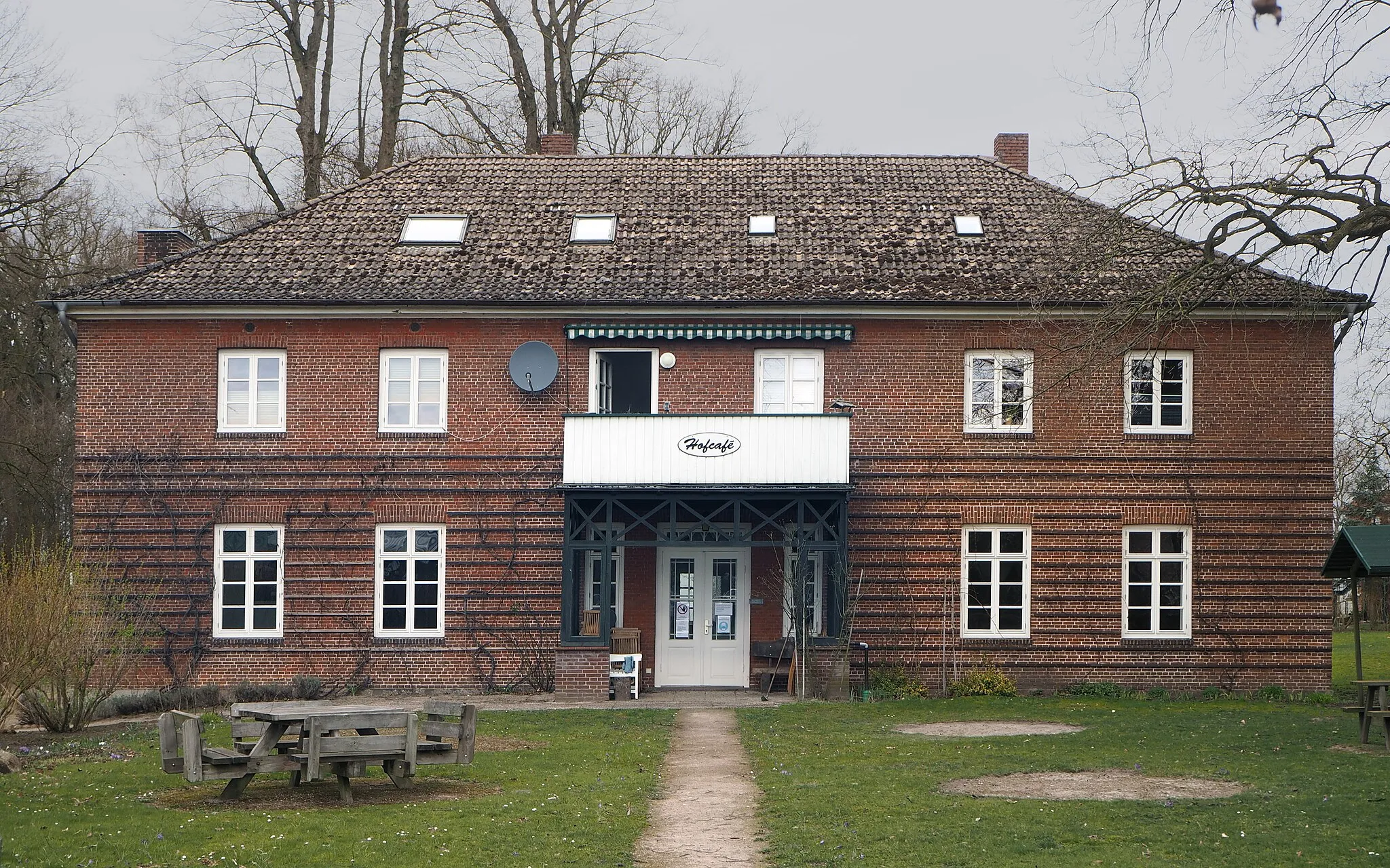 Photo showing: Appen (Schleswig-Holstein) – Schäferhofweg 34 – Nebengebäude mit Hofcafé
– Das Foto zeigt das Nebengebäude der Arbeiterkolonie Schäferhof, welches das Hofcafé beherbergt. Das zweigeschossige, fünfachsige Backsteinhaus trägt ein flaches Walmdach. In der Mitte des symmetrisch angelegten Bauwerks befindet sich der von einem Balkon überdachte Eingang. Die zweiflügelige Eingangstür mit Oberlicht wird von zwei schmalen Fenstern flankiert.