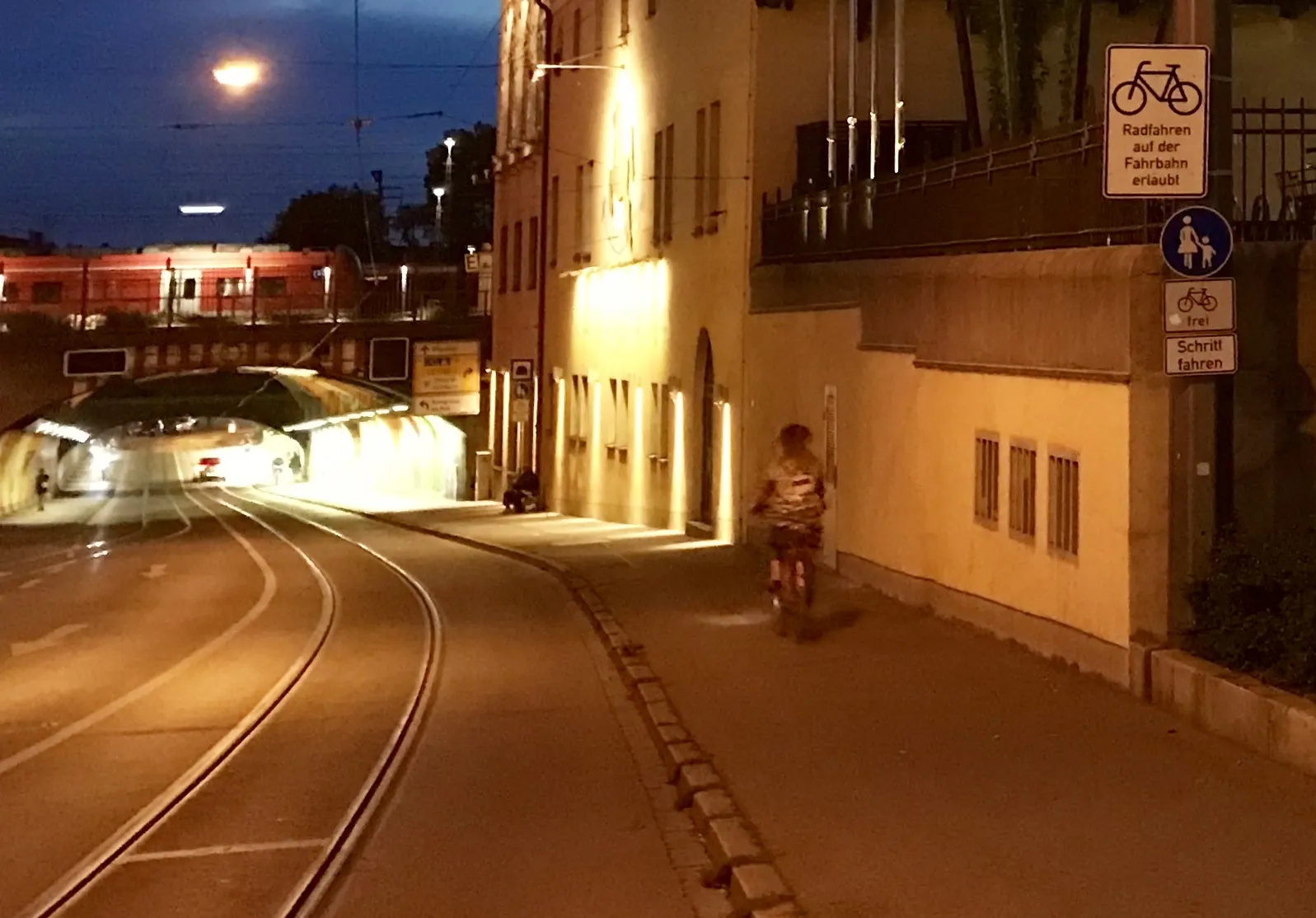 Photo showing: Cycling in Bavaria, street with tram-line-tracks under railway line near Augsburg main station with some people walking on the sidewalk. Building of a brewery (Riegele) in blue hour colors. At that time (2019, evening) not much traffic with cars.