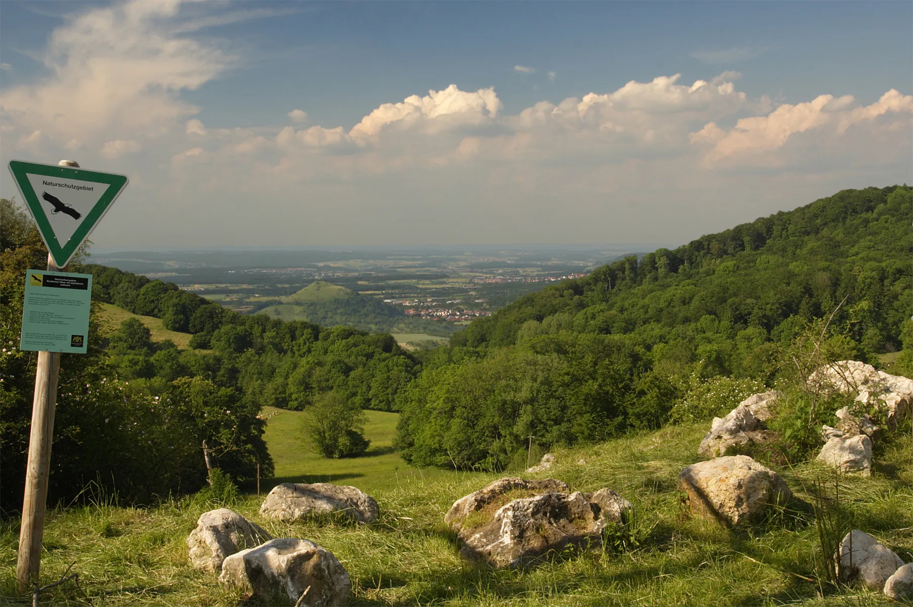 Photo showing: Naturschutzgebiet „Randecker Maar mit Zipfelbachschlucht“. The Randecker Maar used to be a crater lake. The erosive reverse move of the Alb plateau cut the crater in Pleistocene, openening it and emptying the lake. The remaining ground turned out to be a very rich site of fossils in exceptional good state of preservation. Over time the drainage of several karst springs formed a sinking creek with a valuable fauna and flora The crater is one of the volcanos within the Urach-Kirchheimer Vulkangebiet]]. It is a valuable Geotope.
There is evidence of 356 volcanos of the type “Schwäbischer Vulkan”, all extinct in Upper Miocene.
The mountain in the plane is the volcano and nature reserve Limburg.