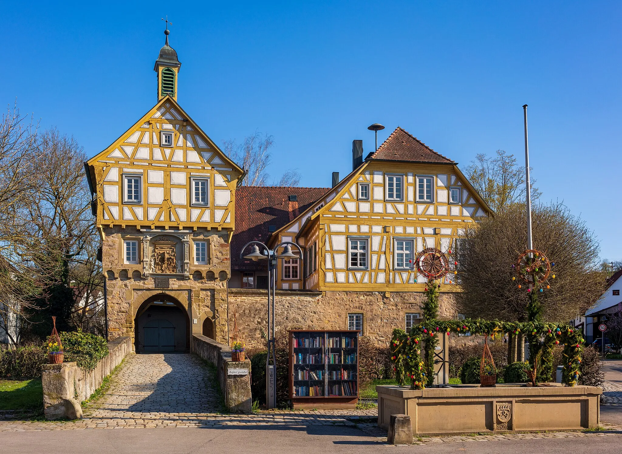 Photo showing: Steinheim an der Murr, Germany, district Höpfigheim: the so-called Schlössle (“little castle”), built in the middle ages as a moated castle and altered in the 16th century to a timber-framed manor house. View from north-northeast. At the right in the foreground the village well from 1984, here decorated as an easter fountain. Hint: The flagpole at the right was really leaning, and the walls and beams of the Schlösschen are not perfectly straight, too, in reality.
