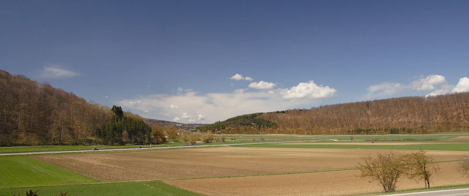 Photo showing: Valley of river Brenz. Todays upper Brenz (hidden beyond the road)  is a poor danubian rivulet. Rivulet and valley beyond it are a strictly protected nature reserve, the visible part of the valley is a protected landscape (Landschaftschutz). The 600m wide valley, ca. 3,5km south of its karst springs (town Königsbronn in the back) cannot be eroded by its contemporary poor rivulet. River Brenz is one of the oldest rivers of Southwest Germany. This is backed by 20 Million years old ”Ochsenberg Schotter”, (river gravel) in a village high above the valley. The catchment area of a pleistocene Brenz reached far beyond the northern border of the Swabian Alb.
The main water devide, now dividing the springs of river Kocher (to the N, river Neckar) and those of river Brenz (to the S, river Danube), occured when the power of water erosion turned to be less than the uplift of the Swabian Alb.