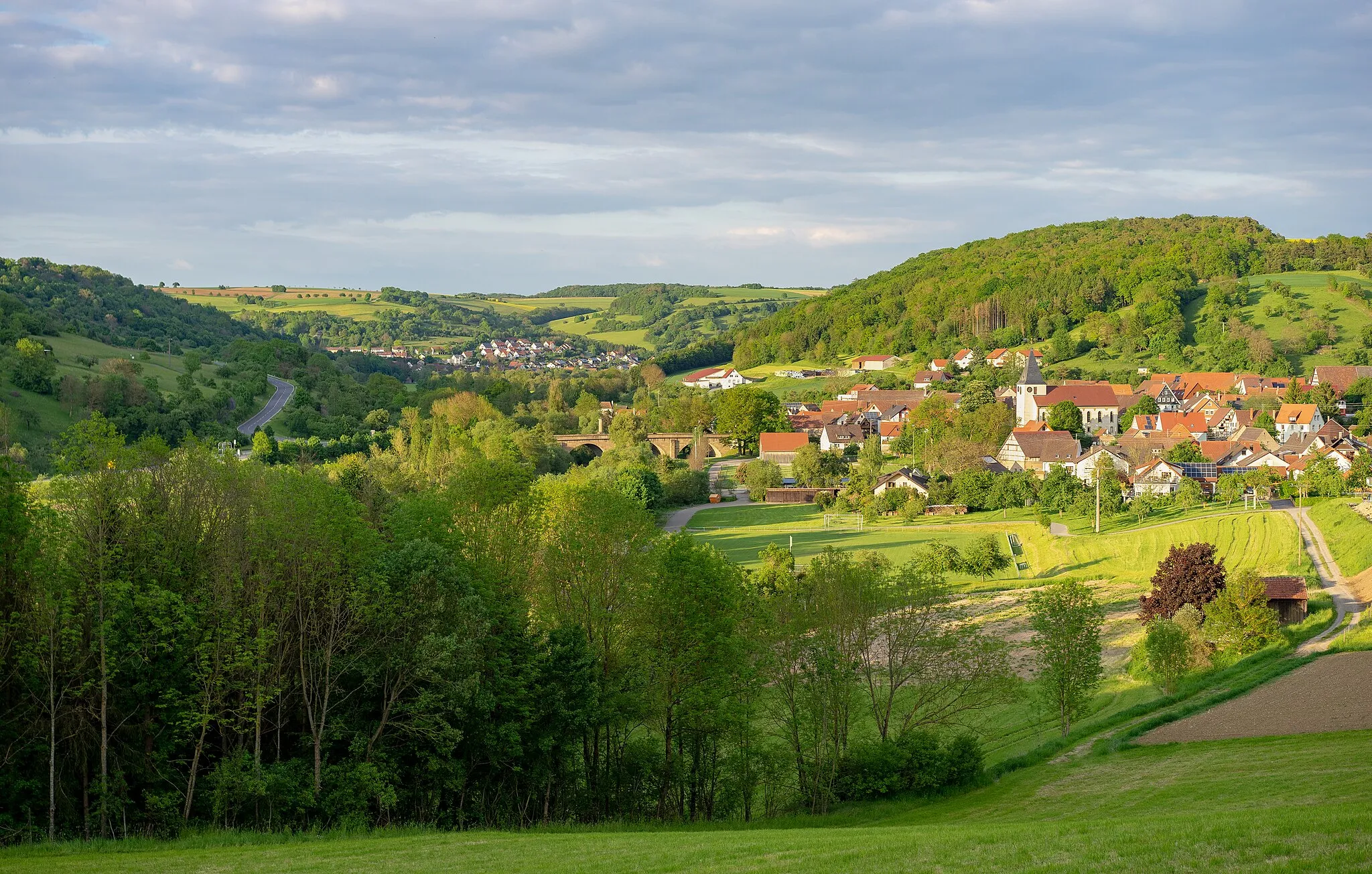 Photo showing: Landschaftsschutzgebiet „Jagsttal mit Nebentälern und angrenzenden Gebieten zwischen Kreisgrenze Schwäbisch Hall und Gemeindegrenze Krautheim/Schöntal“ (Schutzgebiets-Nr. 1.26.029):
Blick von Westen auf das zu Dörzbach gehörige Dorf Hohebach in der Abendsonne. Die Ansicht zeigt gut die Lage des Dorfes zwischen den Hängen des Jagsttals (Landschaftsschutzgebiet). In der Bildmitte die historische Jagstbrücke. Im Hintergrund links das nächste Dorf, Ailringen.