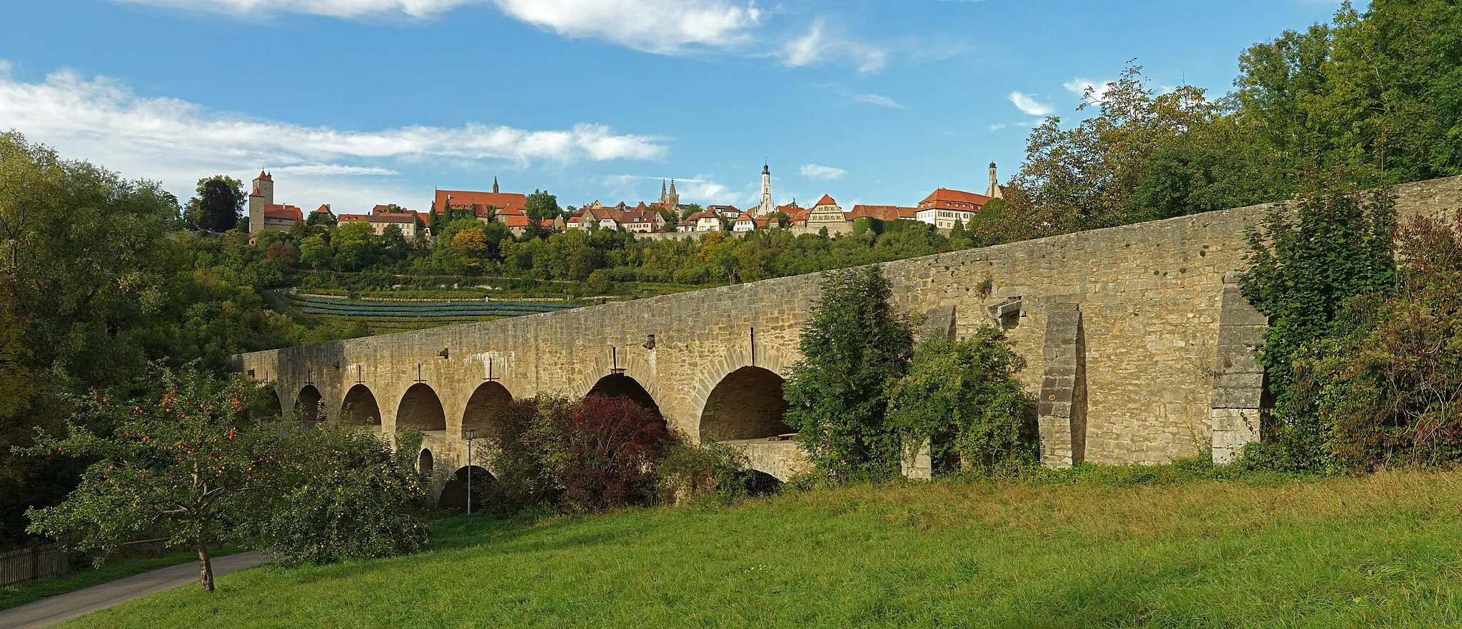 Photo showing: Die Tauberbrücke Rothenburg ob der Tauber ist eine historische Straßenbrücke, die die Tauber überspannt. Die 123 Meter lange Doppelbrücke wurde wahrscheinlich um 1330 errichtet. Im Zweiten Weltkrieg wurde die Brücke gesprengt und 1956 wieder aufgebaut. Etwa 70 Meter oberhalb befindet sich die Kleinstadt Rothenburg ob der Tauber mit einer vollständig erhaltenen Stadtmauer. Blickrichtung ungefähr nordwärts.