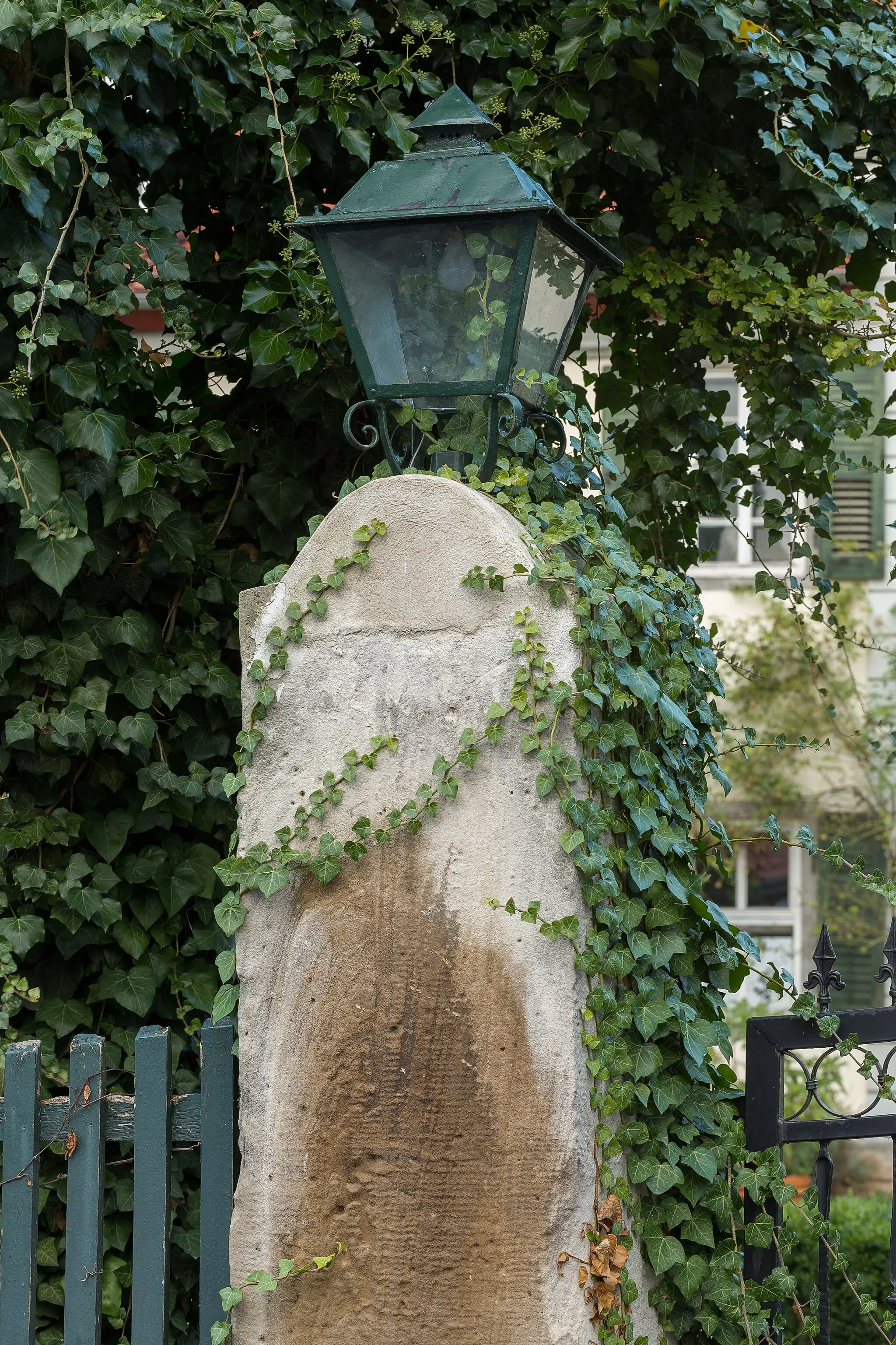 Photo showing: Sandsteinpfosten mit Laterne als Teil des Zauns von Schloss Jestädt. Das Schloss im Hintergrund wird von Efeuranken verdeckt. Im Vordergrund zu erkennen ein Holzzaun (links) sowie ein Teil des eisernen Tores (rechts).