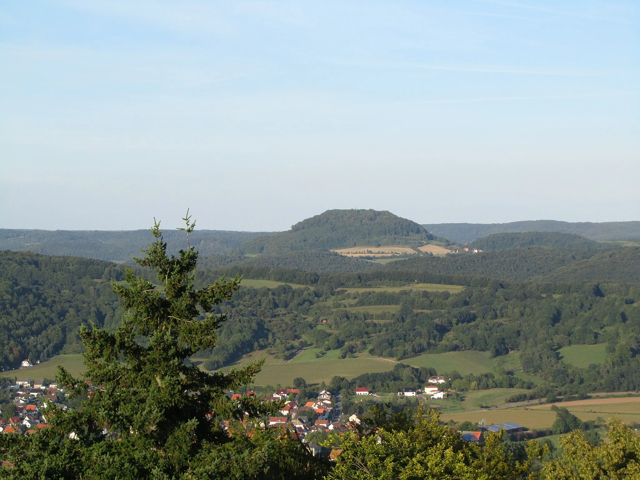 Photo showing: Der Hülfensberg von Südwesten, gesehen vom Bismarckturm auf dem Großen Leuchtberg. Im Werratal die Ortschaft Schwebda, dahinter der Kahlenberg, rechts unterhalb des Hülfensberges ein Teil von Bebendorf, im Hintergrund die Höhen des Obereichsfeldes (links der Westerwald und rechts der Uhlenstein)