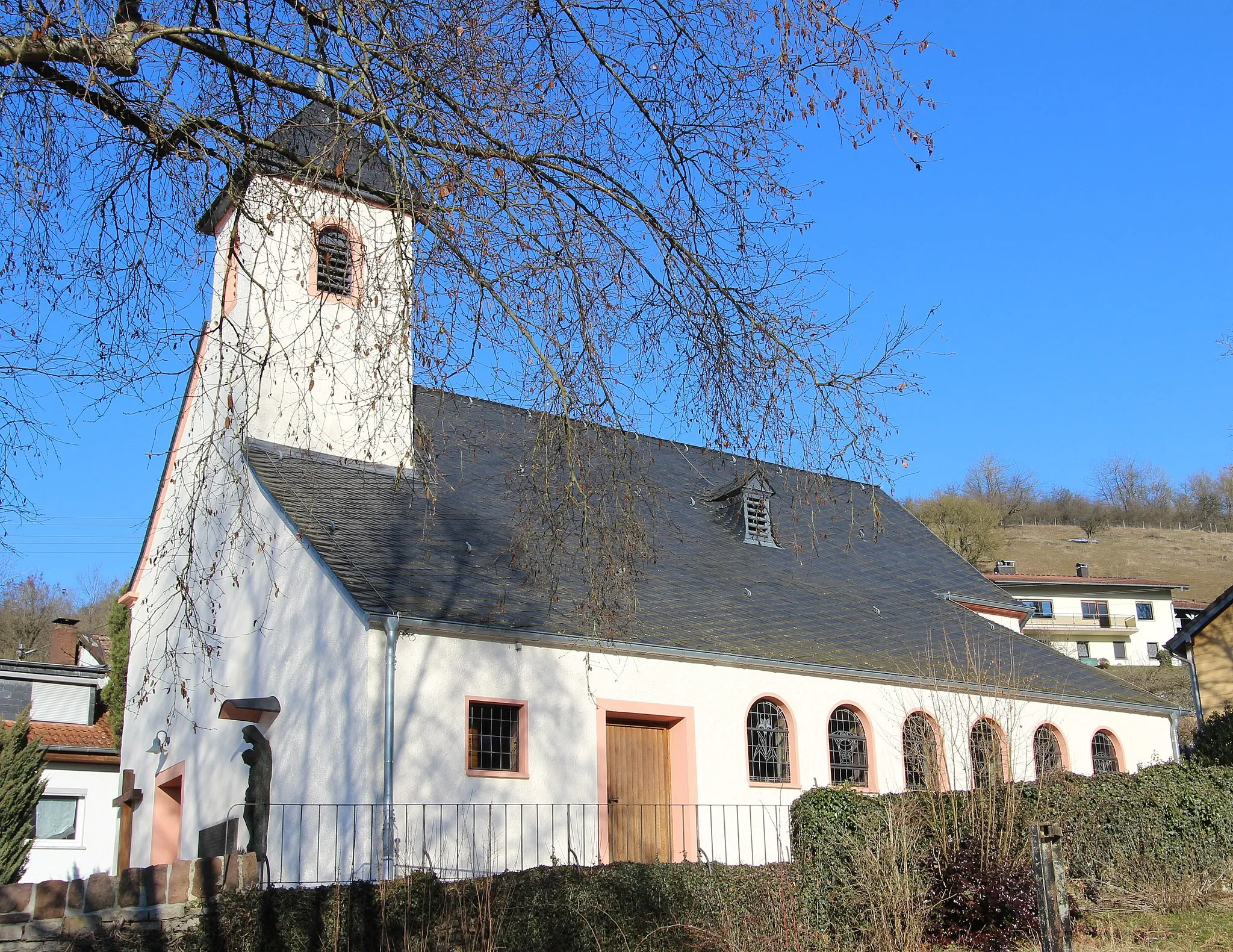 Photo showing: 54636 Hüttingen an der Kyll; Hauptstraße. Westturm (Untergeschoss spätmittelalterlich) und Schiff barock, 1777, 1959/60 Erweiterung zu zweischiffiger Anlage; Kirchhof im Süden mit Kalksteinmauer. Aufnahme von 2017.