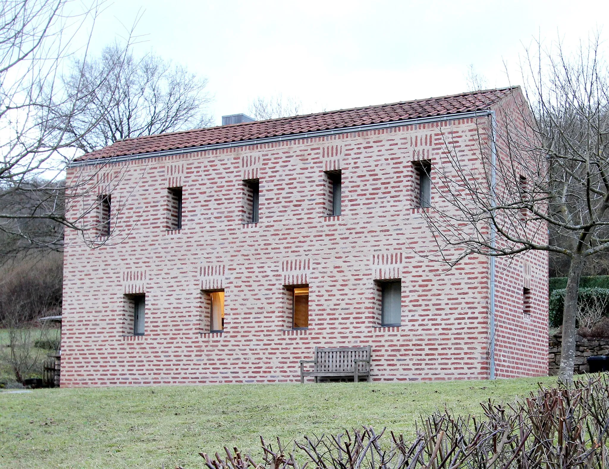 Photo showing: 54636 Hüttingen an der Kyll, Schulstraße 33. Kleines Wohnhaus aus Backsteinaus dem Jahre 2006. Erbaut wurde es vom Architekten Prof. Nikolaus Bienefeld aus Swisttal-Odendorf. Auf der Rückseite ist das Gelände abgegraben und entwickelt mithilfe der Stützmauer einen kleinen Hof. Die rückseitige Glasfront füllt den Innenraum mit viel Tageslicht und bezieht den Hof mit in das Haus ein. Das nicht unterkellerte, zweigeschossige Gebäude verfügt über eine Fußbodenheizung (Erdwärme) sowie über einen offenen Kamin. Aufnahme von 2017.