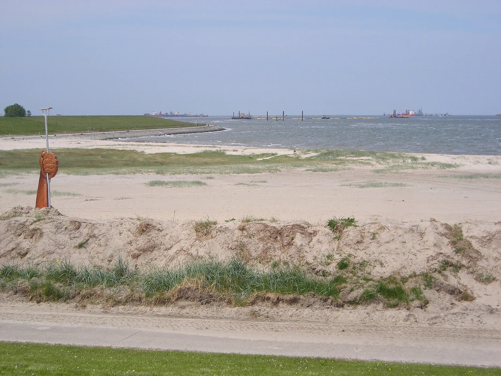 Photo showing: JadeWeserPort construction site at Wilhelmshaven, Germany. In the foreground the Geniusstrand beach with a sign of protest against the JadeWeserPort project (left). In the background floating sand flushing tubes (center) and oil loading terminals (left and right)