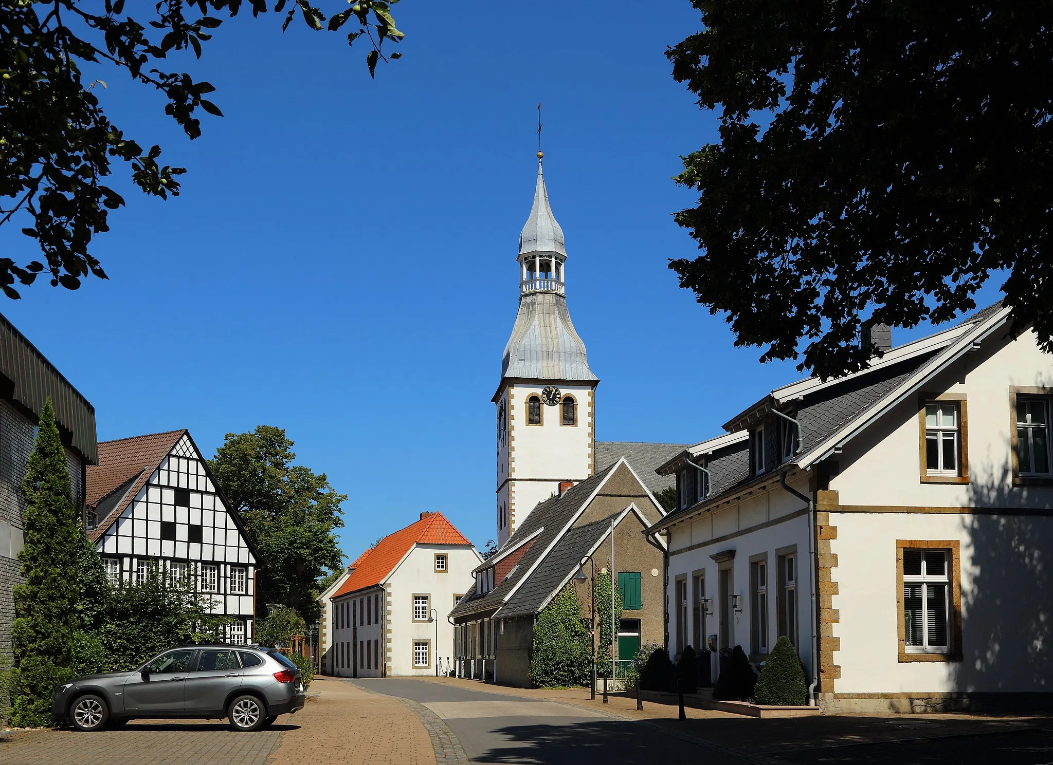 Photo showing: Die Brenninkmeyerstraße im Ortskern von Hopsten, Kreis Steinfurt, Nordrhein-Westfalen, Deutschland. Das frühere Kopfsteinpflaster wurde 2013 im Zuge einer Umgestaltung eines Abschnitts der Straße durch Betonsteinpflaster ersetzt. Dies soll die Barrierefreiheit dort erhöhen. Im Hintergrund ragt der Turm der Pfarrkirche St. Georg auf.