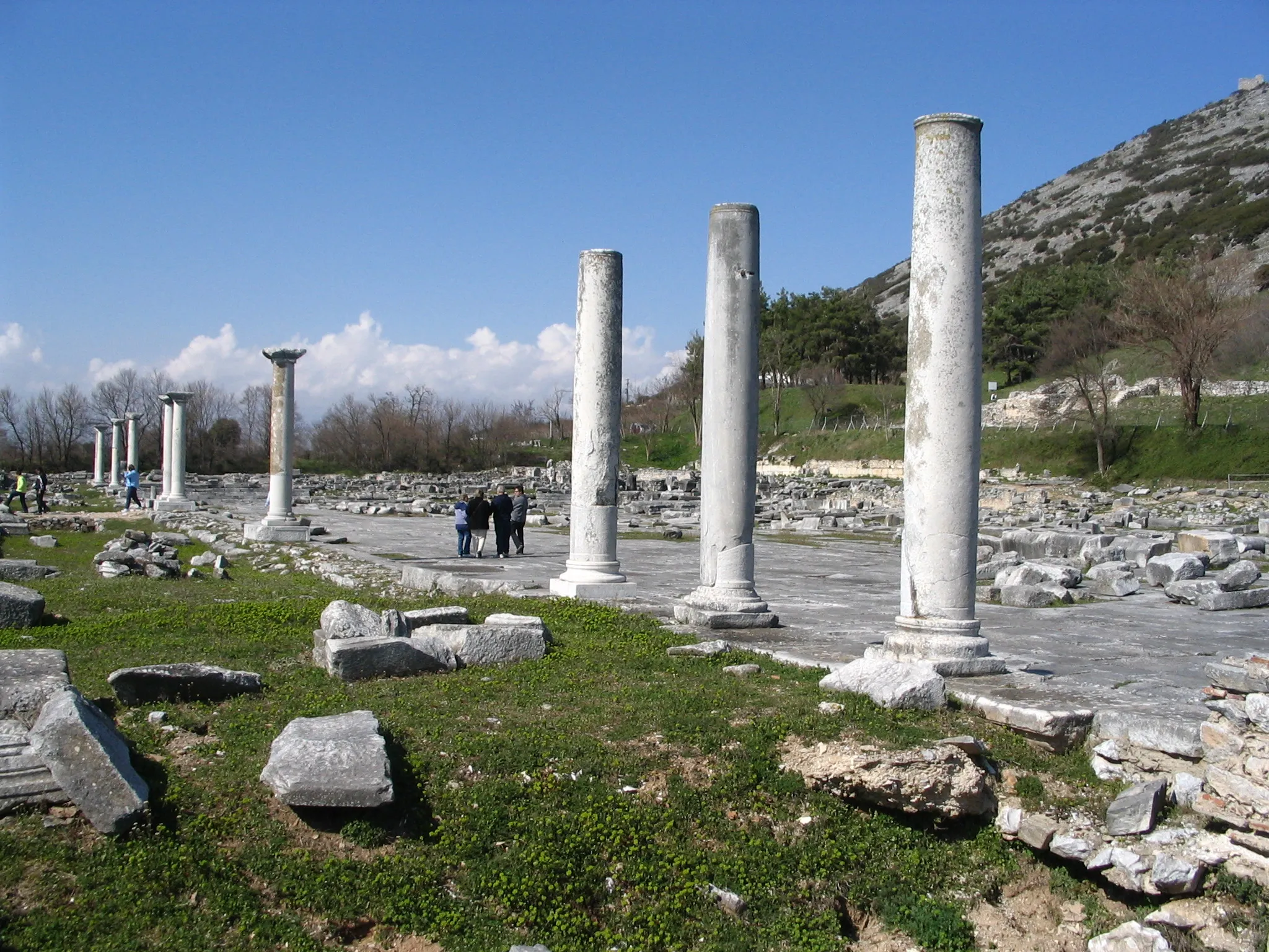 Photo showing: Ruins of ancient Philippi, looking NW along the southern portico (stoa) of the city's main marketplace (or agora). A roof would have covered the grassy area along the near side of the line of monumental columns. The broad stone-paved area beyond was the open market area. Along the far (northern) side of this market ran the via Egnatia, the main Roman highway through Macedon. In the distance (at right) is visible the acropolis, or high hill, of the city. The Roman inhabitation spread up much of the hill's SW face.