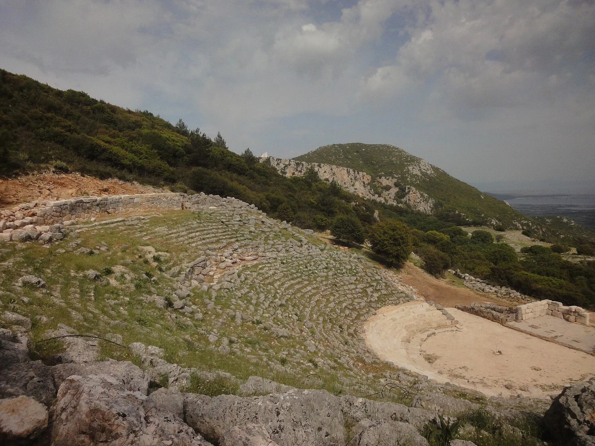 Photo showing: The theatre of ancient Cassope, Preveza, Greece, after the restoration of the first five rows of seats. The w:en:Dance of Zalongo:monument of Zalongo can be seen in the background.