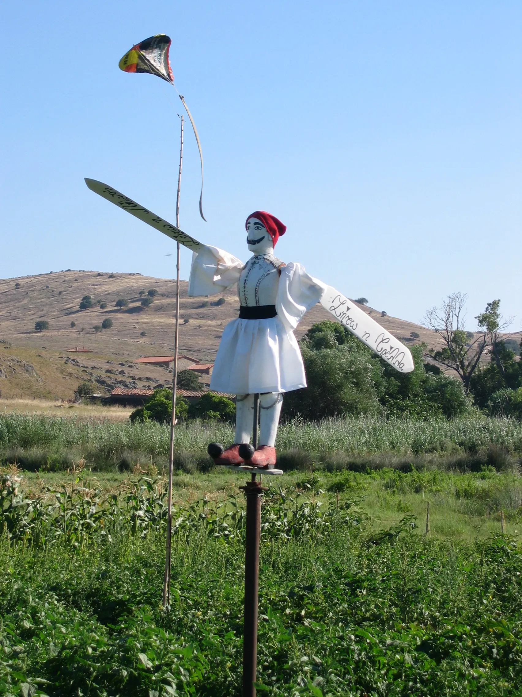 Photo showing: Crops in Greece tend to include scarecrows. This one, on a crop field in the island of Lemnos, Greece, has the form of an Evzonas, a Greek soldier dressed in traditional clothing. Its arms have the shape of oars, so the dummy rotates depending on the wind's direction. In the left arm it is written, in Greek calligraphic script "Long live madness" (Ζήτω η τρέλλα), while in the right arm it is written "Long live Lemnos" (Ζήτω η Λήμνος).