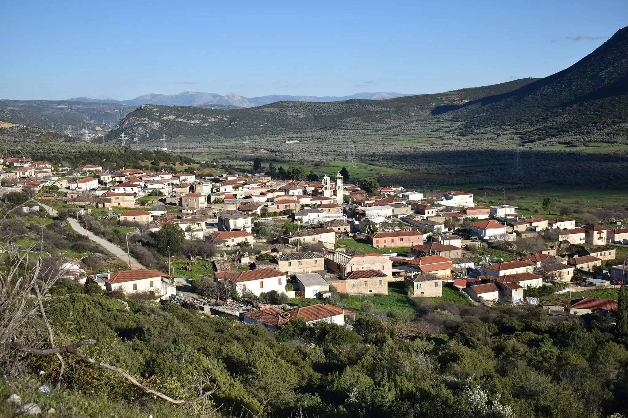 Photo showing: In the centre is the village with the bell towers of the church of the Dormition of the Theotokos. On the upper right lies the plain of the river Inachos filled with olives, and over it rise the slopes of Mount Brachriami.