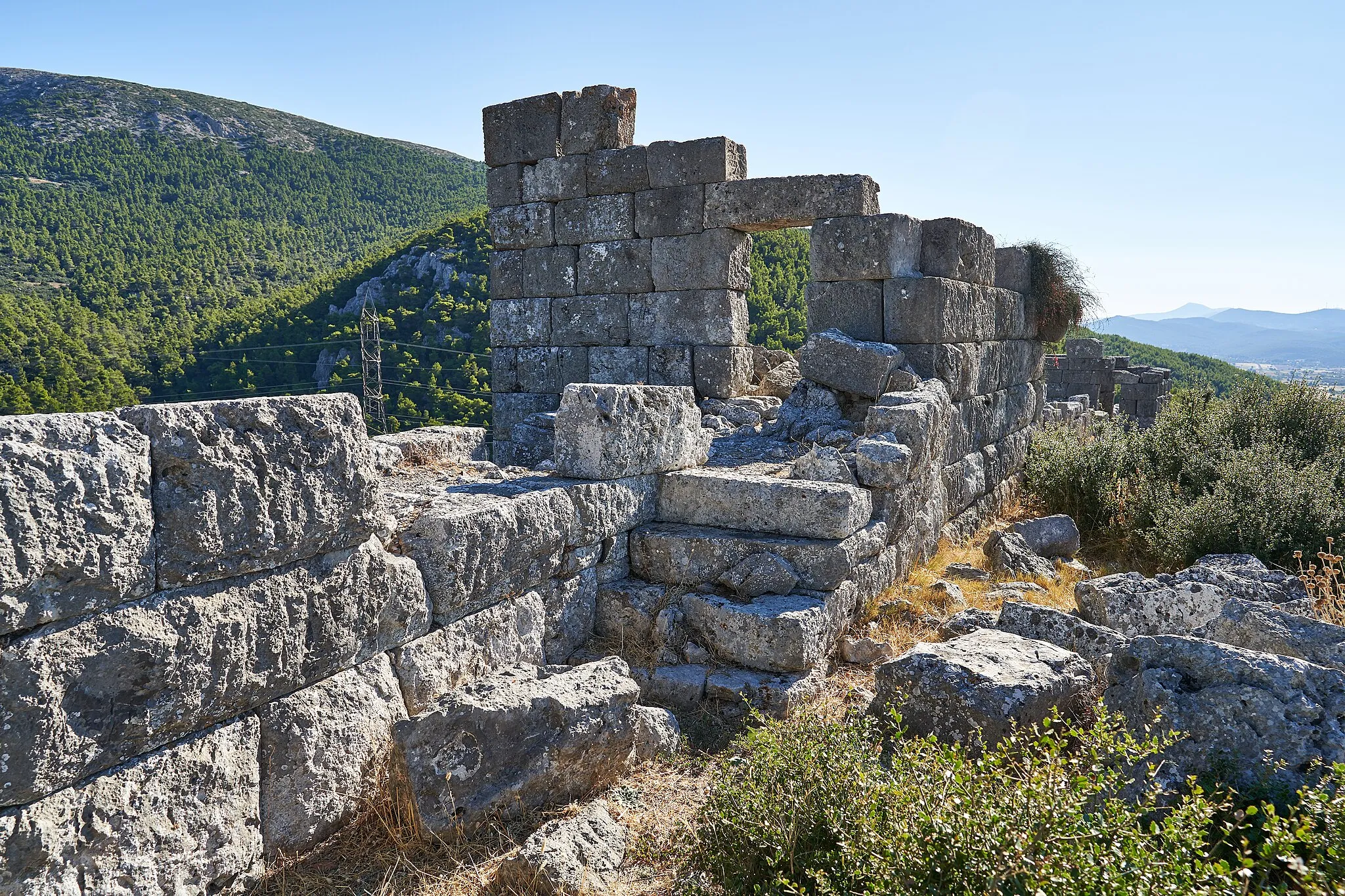 Photo showing: “Fortress of Eleutherai
The fortress of Eleutherai has been built at a strategic position for control of the narrow pass leading from Boeotia to Attica. From this point there is an unbroken view toward the ancient passage to the west and over the plain of Oinoe to the southeast. Its date between 370 and 360 B.C. places it in the Athenian defence system that was evidently constructed along the borders of Attica during the 4th century B.C.
The line of the fortress follows the line of the hilltop, enclosing an area of 300 x 100 metres, with circuit walls 860 metres in length. The vulnerable north side has a strongly constructed wall, preserved to a height of some 4 to 6 metres, with two small sally - ports. It is reinforced by seven square towers, 6 metres along the side, that project from the two faces of of the wall and are connected by a curtain wall with a parapet - walk. Two gateways provide entrance to the fortress: the main, double gateway is at the west above the control -road; the secondary entrance, at the southeast side, with a small gateway, connects the fortress with the city of Eleutherai by way of the south slopes of the hill. In addition to the north side, the other sides two are reinforced, but with fewer towers preserved only to a low level. Their masonry is in places polygonal and irregular, evidently because of the terrain.
Preserved in the interior, close to the north side, are the foundations of a building measuring 16 x 11 metres, evidently with two storeys, with polygonal wall construction, a central passageway and a pair of rooms at each side. It is probably to be dated to a phase earlier than the fortress and it will have served as in installation facility for a garrison. It is possible too that it served for billeting even after the fortress was completed. Fragmentary low walls, scattered through the interior area of the fortress, indicate that there were other such buildings as well.
3rd EPHORATE OF PREHISTORIC & CLASSICAL ANTIQUITIES”

Text: Site marker inside the fortress.