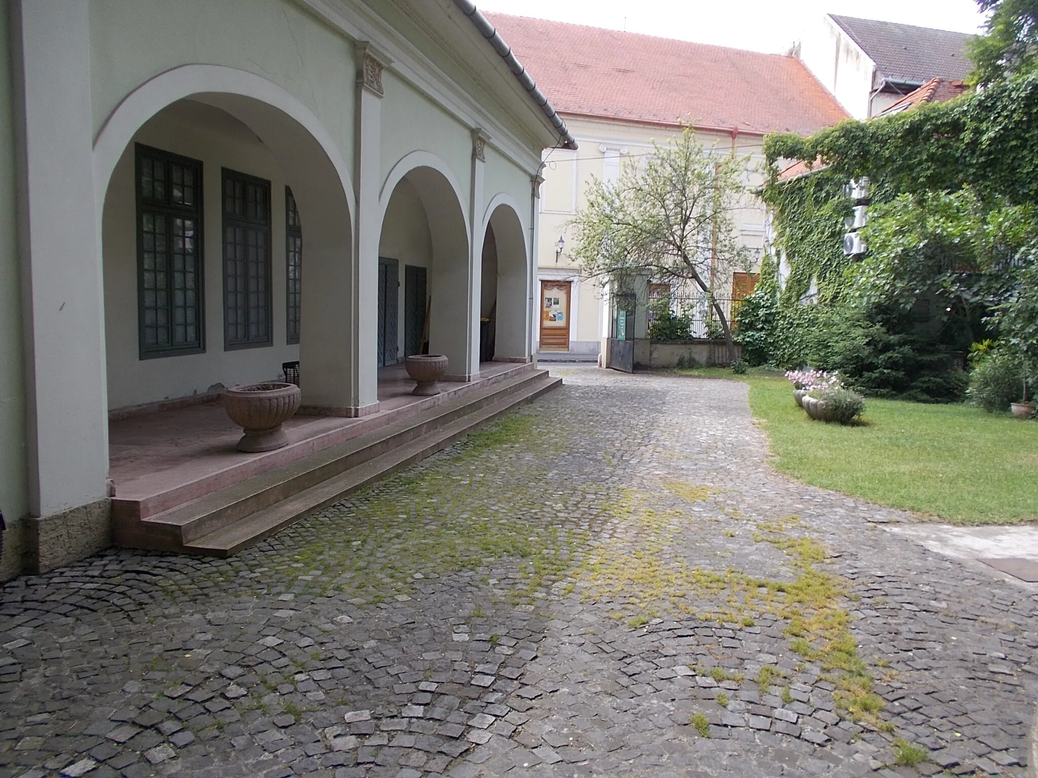 Photo showing: Former Vojnich country house. Since 1985 museum. In 2023 Nagy István Gallery. Built in the 1830s probavly to György Vojnich. Later it became the property of the Herczfeld family, and from the 1920s it became the property of the Grünhut family. Between 1947 and 1952 Gyula Rudnay's painting school operated here, and since 1985 it has been a museum. The building: freestanding, rectangular, ground-floor, gabled roof. At its southeast corner is a circular corner rizalite. On its southern, street façade, it is a central rizalite articulated by pilasters and closed by a tympanum. The pilaster chapters are decorated with Kossuth coats of arms, in the tympanum is the coat of arms of the Vojnich? family. In front of its northern, garden façade, there is a four-column, tympanum portico, and to the west of it is a basket-arched, arcaded corridor.  - 1 Arany János Street (Oroszlán Street corner), Baja, Bács-Kiskun County, Hungary.