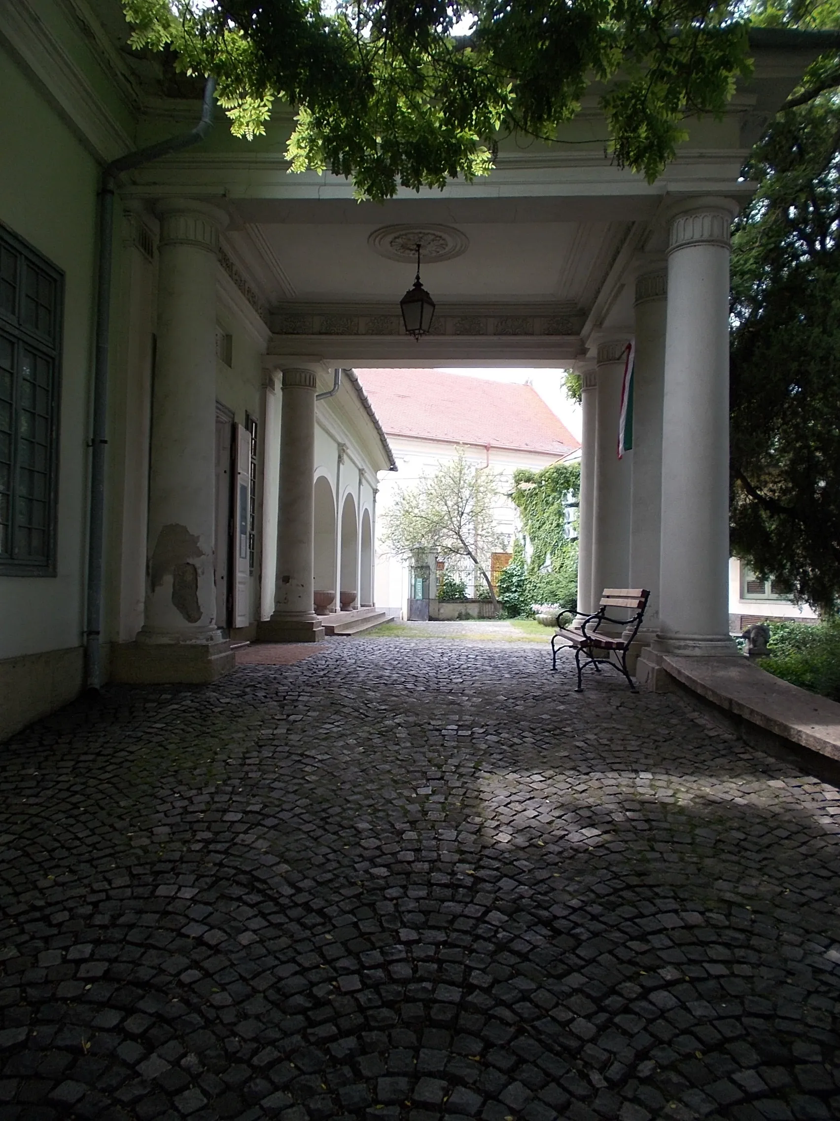 Photo showing: Former Vojnich country house. Since 1985 museum. In 2023 Nagy István Gallery. Built in the 1830s probavly to György Vojnich. Later it became the property of the Herczfeld family, and from the 1920s it became the property of the Grünhut family. Between 1947 and 1952 Gyula Rudnay's painting school operated here, and since 1985 it has been a museum. The building: freestanding, rectangular, ground-floor, gabled roof. At its southeast corner is a circular corner rizalite. On its southern, street façade, it is a central rizalite articulated by pilasters and closed by a tympanum. The pilaster chapters are decorated with Kossuth coats of arms, in the tympanum is the coat of arms of the Vojnich? family. In front of its northern, garden façade, there is a four-column, tympanum portico, and to the west of it is a basket-arched, arcaded corridor. - 1 Arany János Street (Oroszlán Street corner), Baja, Bács-Kiskun County, Hungary.