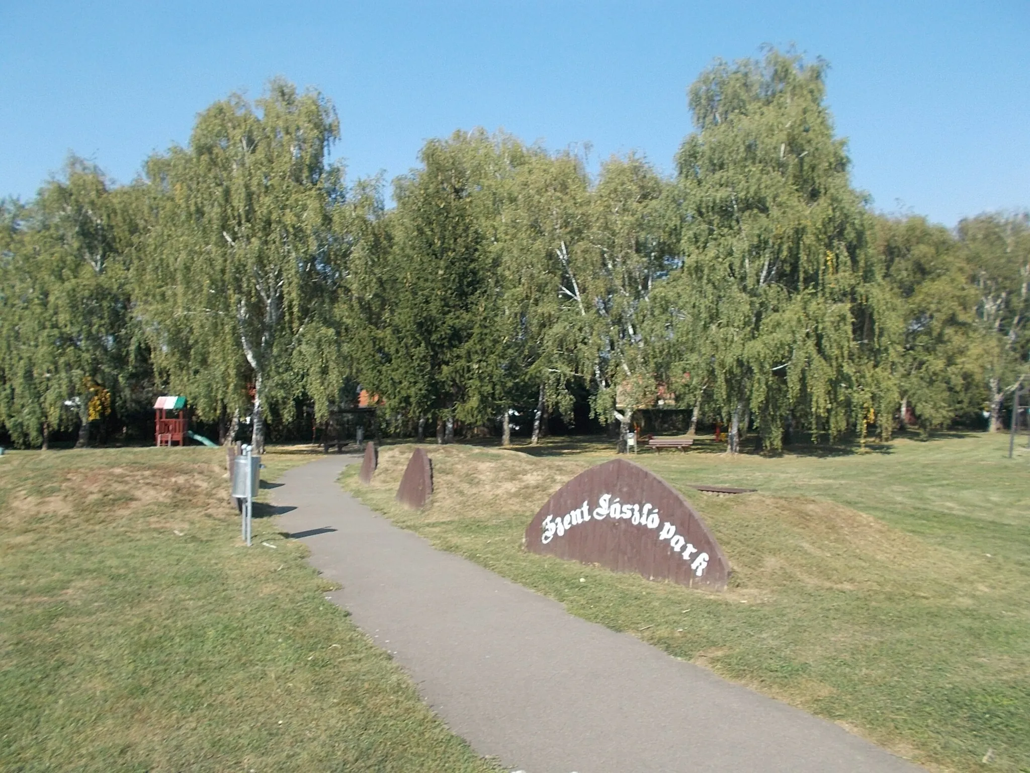 Photo showing: : Szent László (Saint Ladislaus) Park, name sign, and the hills are perhaps a mountain bike track? in the background between the trees a playground. - Gyöngyvirág körút, Kertváros (Garden City), Dombóvár, Tolna County, Hungary.