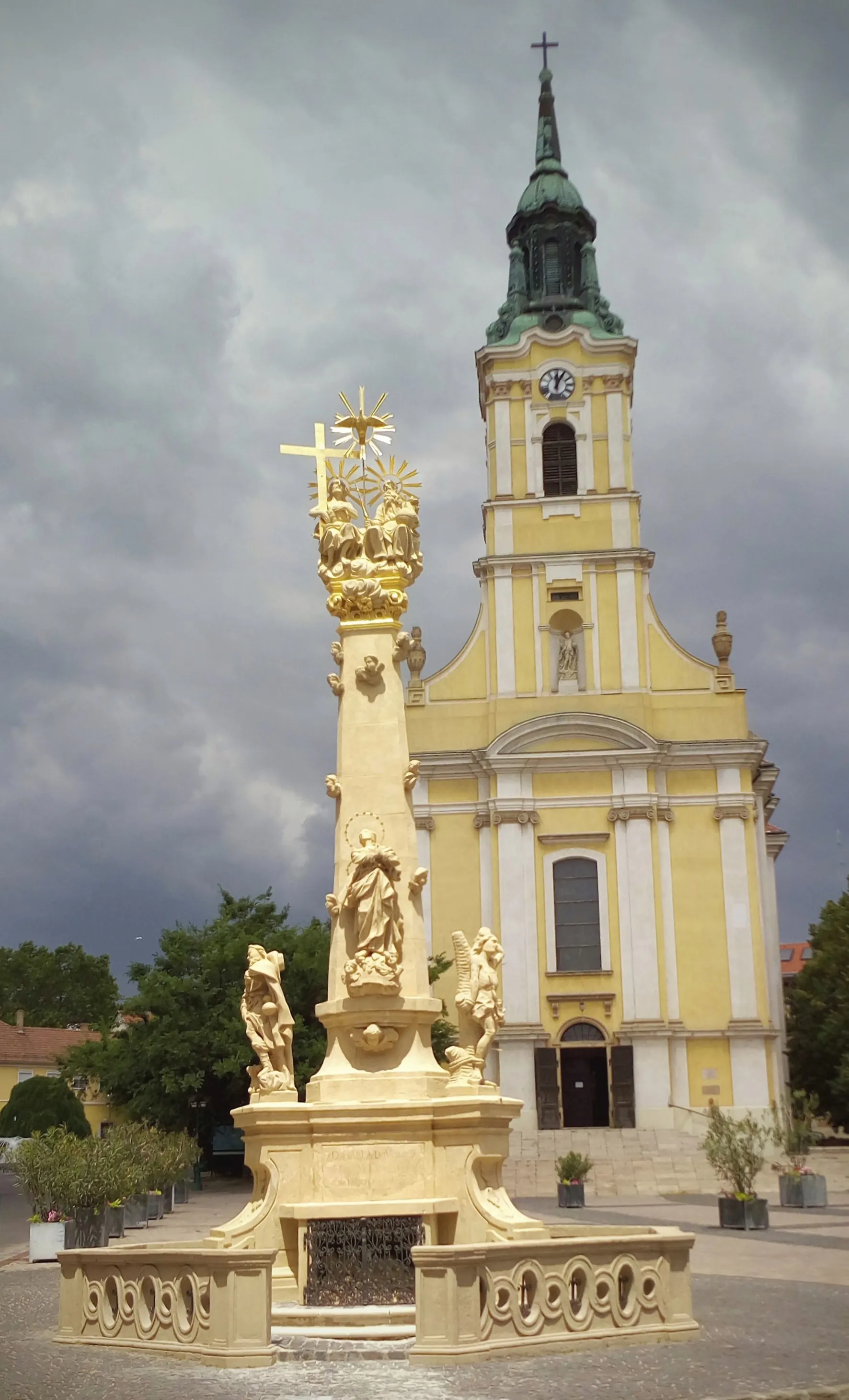 Photo showing: A 2017-ben felújított Szentháromság szobor a copf stílusú katolikus Urunk Mennybemenetele templommal a háttérben.