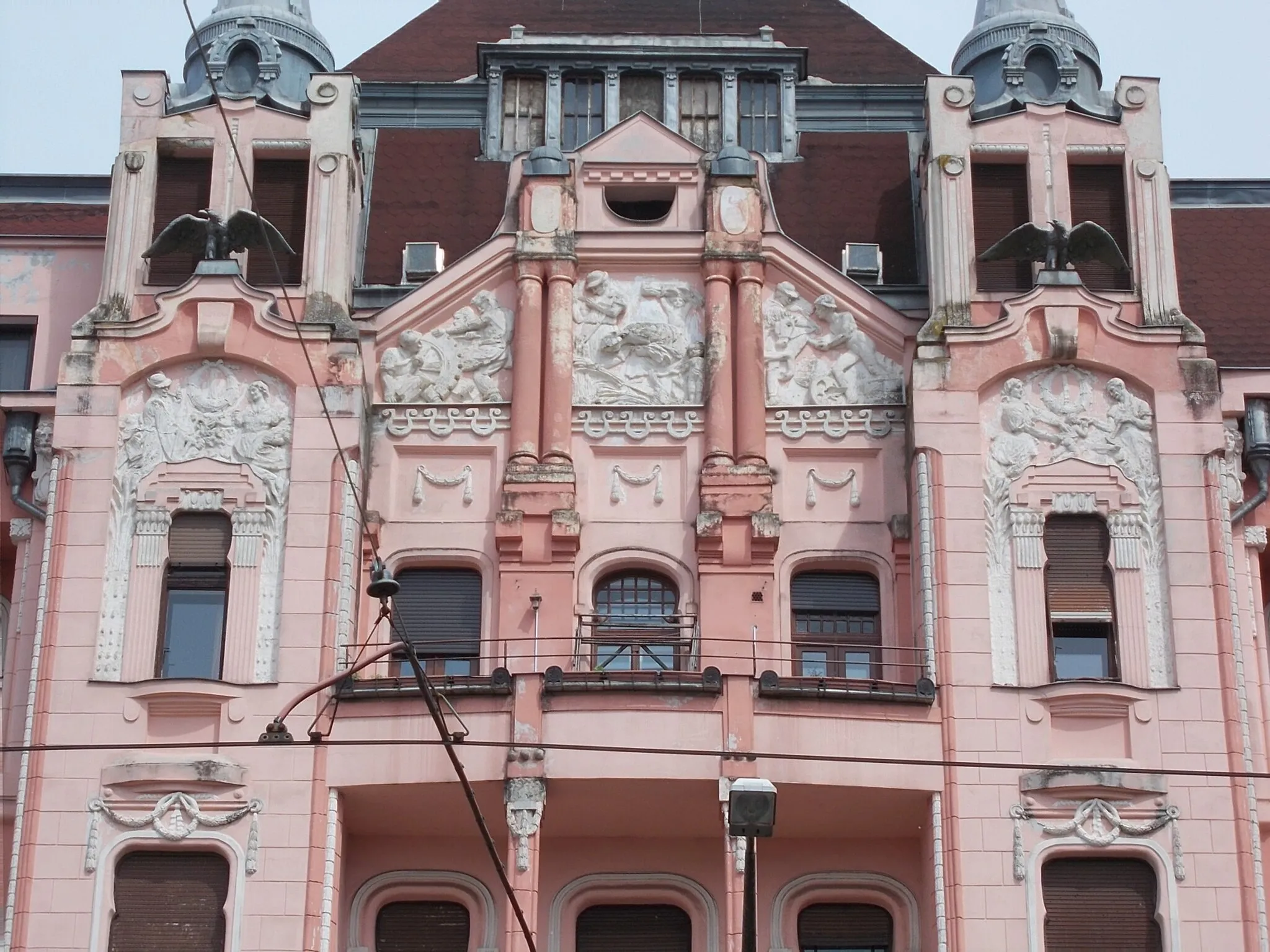 Photo showing: HQ of the former Debrecen First Savings Bank, west facade, mid part, stuccos and bird statues. Buil in 1912 Planned by Kálmán Rimanóczy Jr.. Construction contractors: István Tóth, Szilágyi and Pavlovits. Facade allegorical sculptures related to agriculture, industry and commerce  and the nude male stuccos are Sándor Somogyi works ). There is couple bronze bird statues (Turul?). Inside coulmn capatáls made from Zsolnay ceramic. In addition to the premises of the Savings Bank, there were shops on the ground floor, on the Kossuth street side there were originally an officer's casino, in the cellar there was a beer hall and restaurant, on the floors there were seventeen luxury rental apartments. - 22-24 Piac Street, Inner City,Debrecen, Hajdú-Bihar County, Hungary.