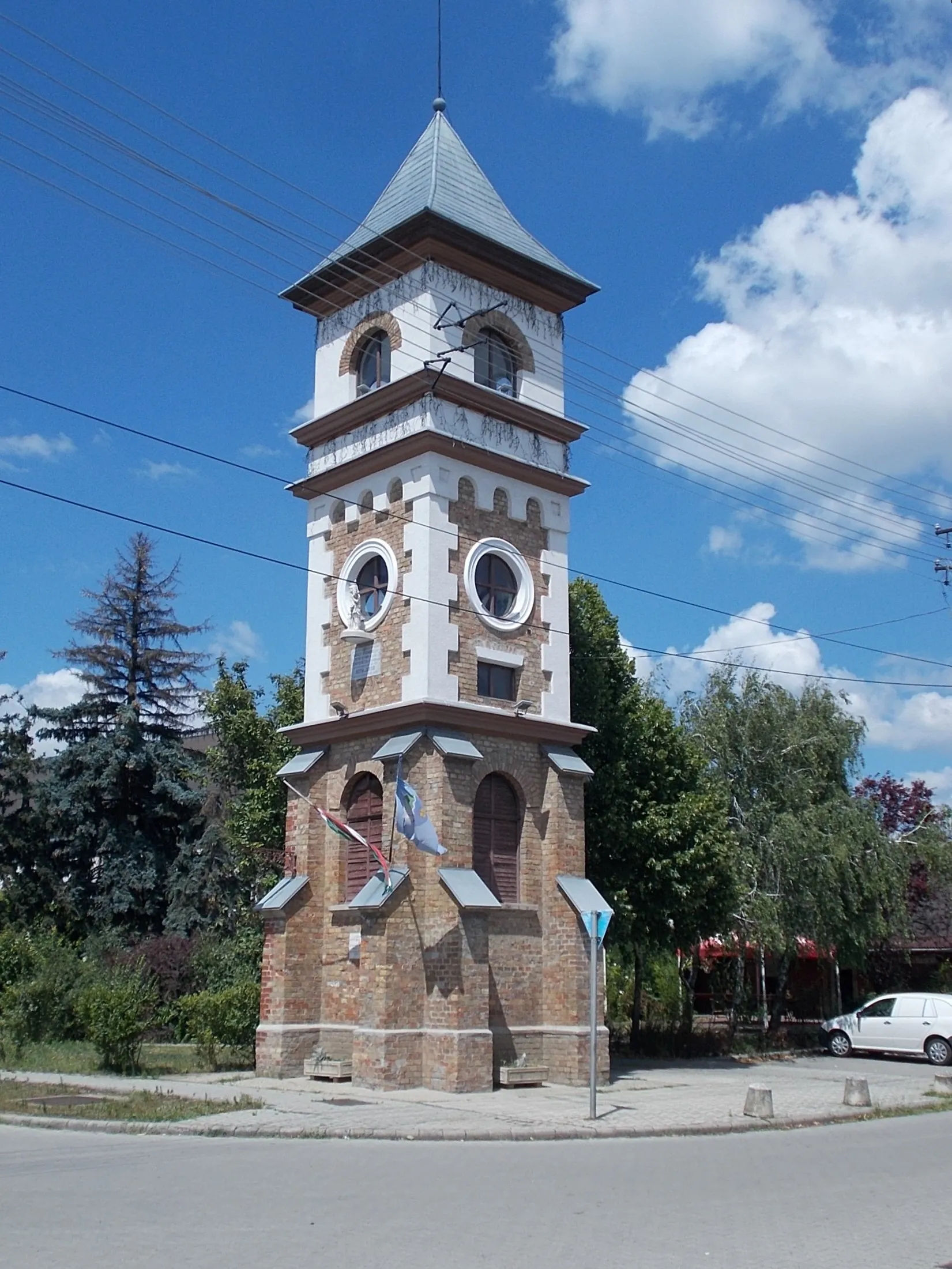 Photo showing: Local grade listed fire lookout tower. It was built in 1903 to protect the village's then mostly thatched-roofed houses in historicist style. It was renovated in 1978, 1998 and 2015. In front of a round window in the middle of the tower you can see the statue of St. Florian, the patron saint of firefighters. Under the statue is a memorial plaque announces that the fire tower was: ~"It was built by the village in 1903 during the official office of judge Zsigmond Károly, notary István Gerő, and Commander-in-Chief Dezső Tschepen Ö.T.E. by local entrepreneur Jakab Róth." /note: Ö.T.E.=abbr.: Volunteer fire department /. - The inscription on the memorial plaque on the facade ground floor part: ~"The fire tower was restored by the craftsmen of the village in 1978 in honor of the 30th anniversary of the establishment of KIOSZ." KIOSZ=abbr. National Organization of Craftsmen. - Szent István Road and Táncsics Mihály Street corner, Ercsi, Fejér County, Hungary.