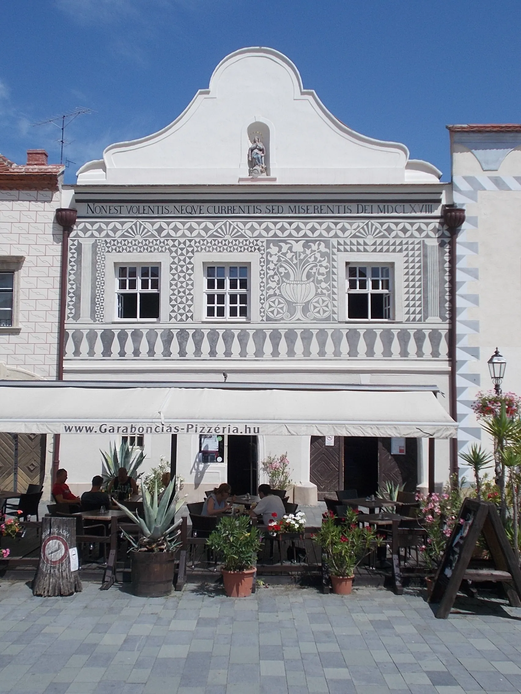 Photo showing: Renaissance, 16th century building with sgraffito (around 1560). The courtyard wing built in the 18th and 19th century. Immaculata niche statue (1668 /other source 1720/ baroque limestone). Garabonciás Café sign by János Lehocky (1974 abstract iron, copper) can be see on its facade. The former Garabonciás Café, now Garabonciás Pizzeria. Ubnder ledge Latin inscription: "Non Est Volentis, Nequue Currentis Sed Miserentis dei MDCLXVIII" - 7 Jurisics Square, Kőszeg, Vas County, Hungary