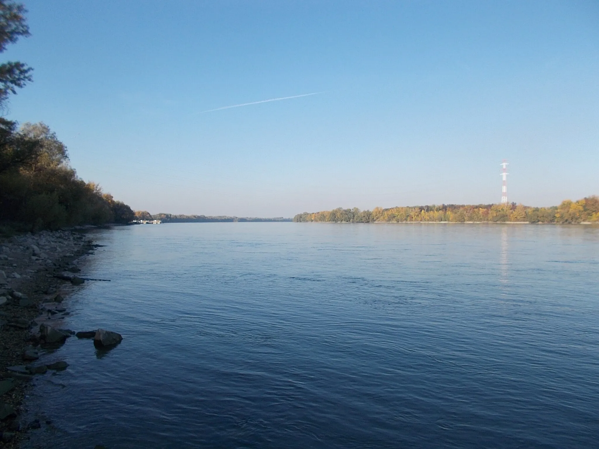 Photo showing: : View to north. On left at the former ship station are barges. On right at Szalkszentmárton ferry 'departure zone' is a high electric pylon (part of the 120 kV overhead power line crossing of the Danube river at Dunaújváros). Viewed from the east shore of Szalki Island, Dunaújváros, Fejér County, Hungary.